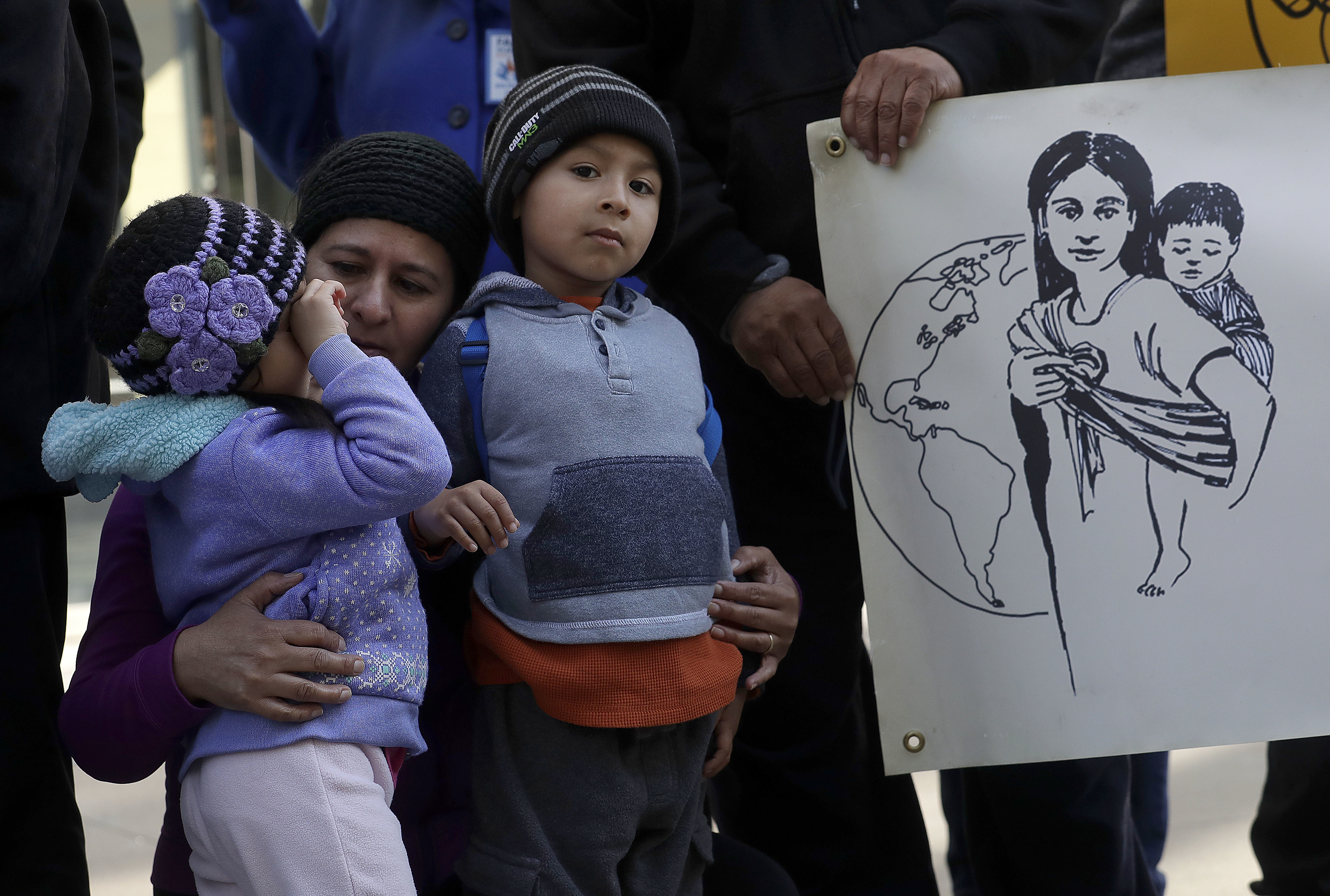 A woman crouches with two children in winter clothing, while a hand holds a poster showing a mother carrying a child with a globe in the background.