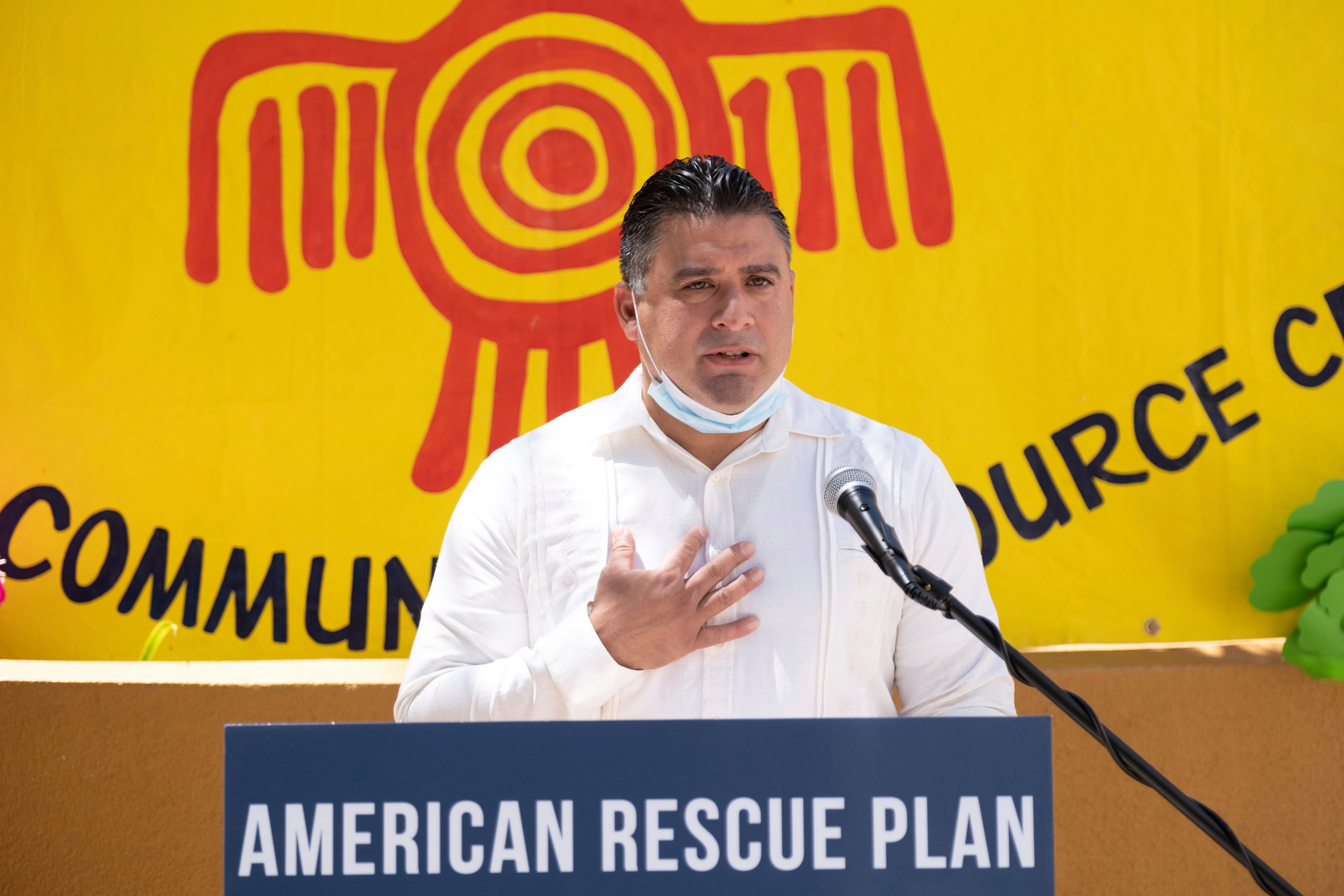 A man in a white shirt speaks at a podium labeled &quot;American Rescue Plan.&quot; He gestures with his hand on his chest, standing before a bright yellow backdrop.