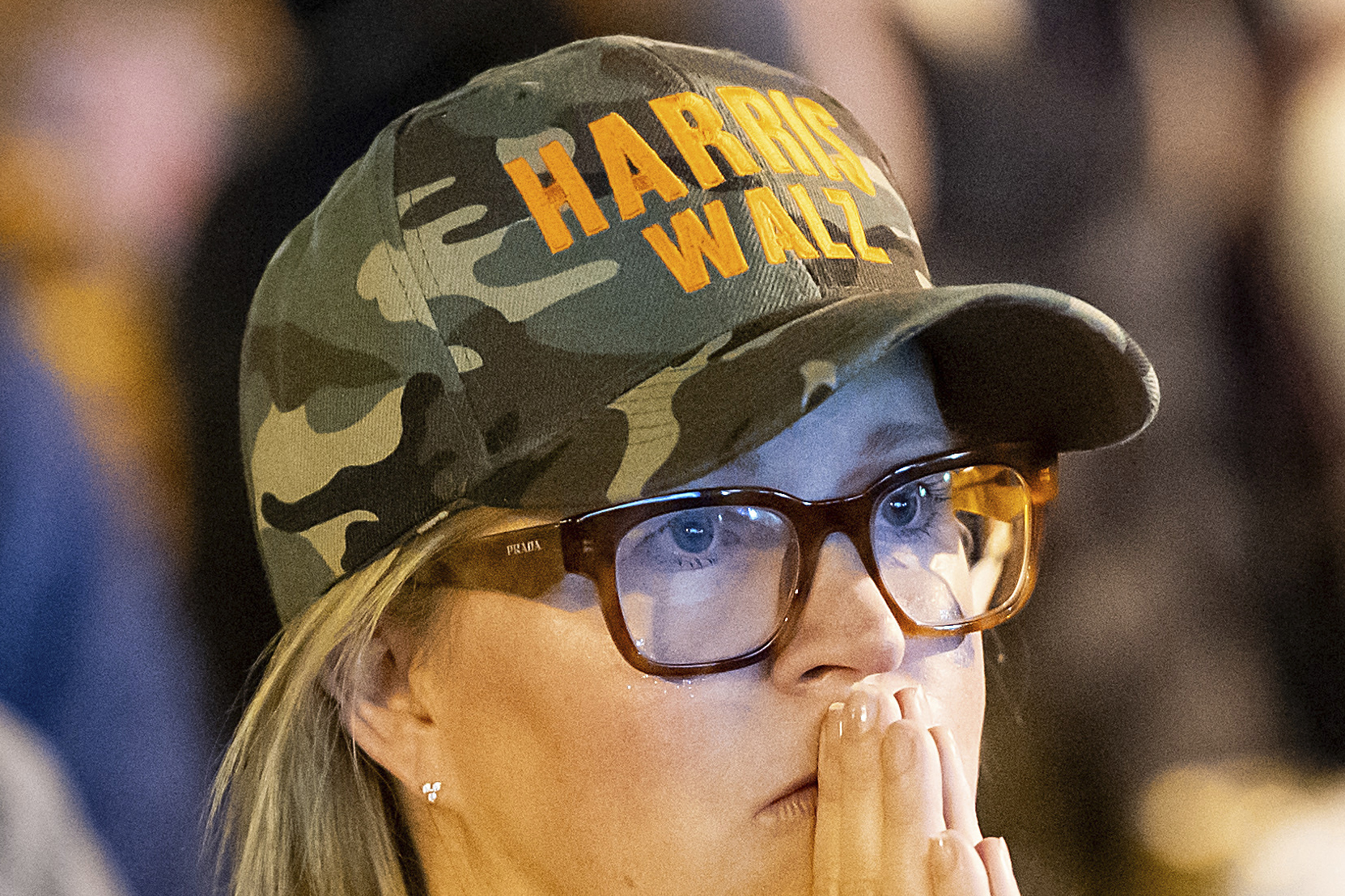 A person wears glasses and a camo hat with &quot;HARRIS WALZ&quot; in orange. They have a thoughtful or concerned expression, with hands partially covering their mouth.