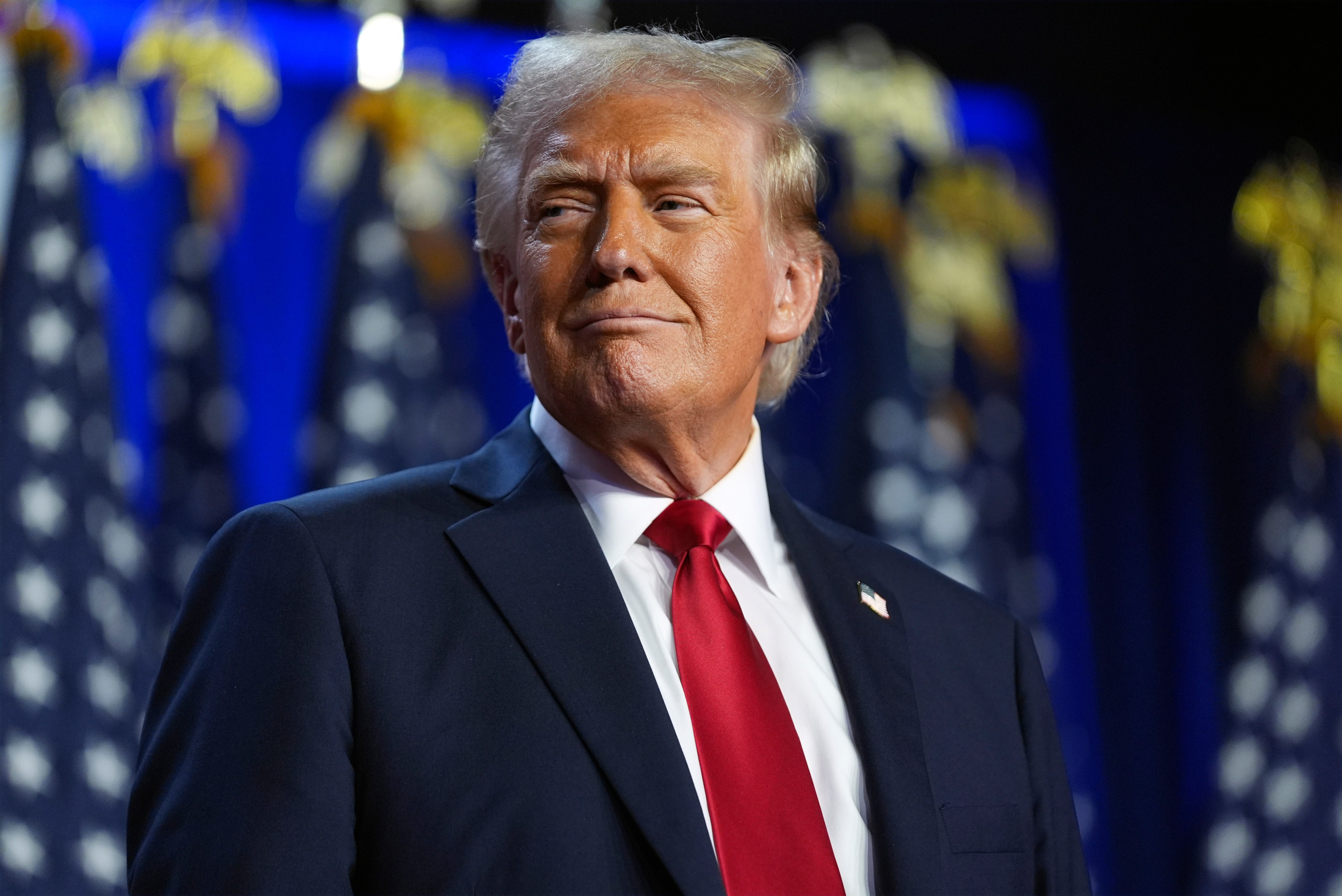 A person with blond hair is wearing a dark suit and red tie, looking to the side with a confident expression. The background shows blurred US flags.