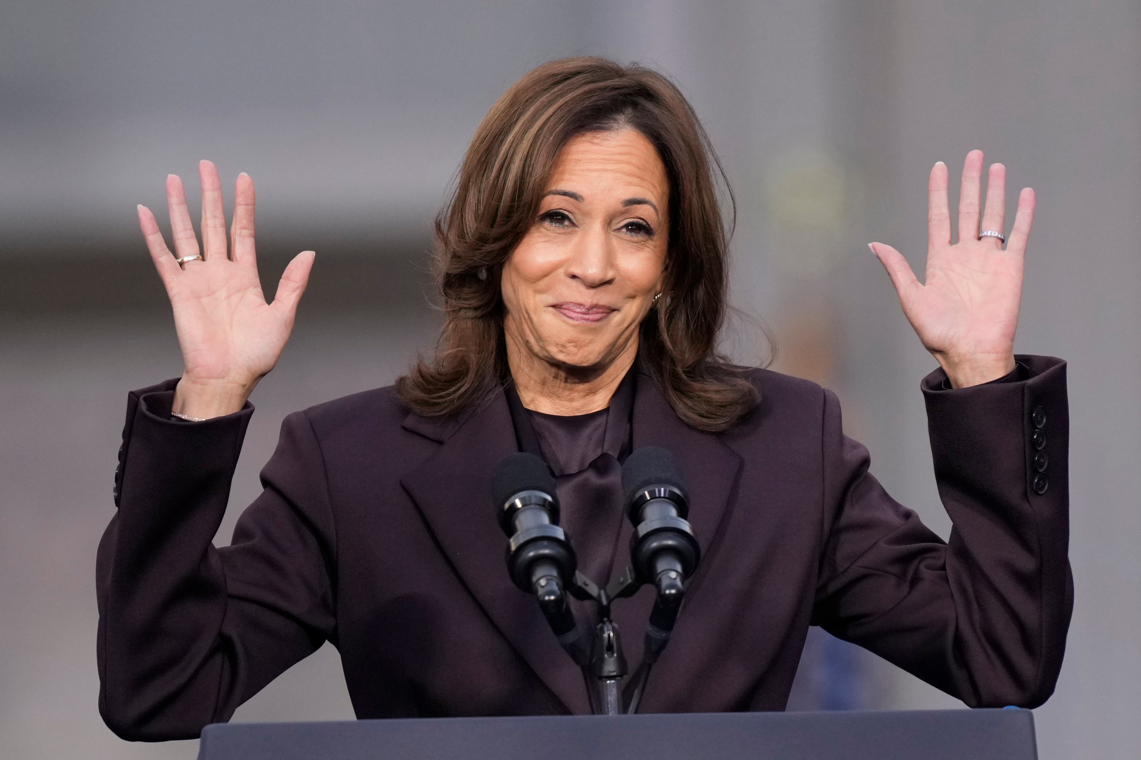 A woman stands at a podium with a microphone, wearing a dark suit, smiling, and raising both hands.