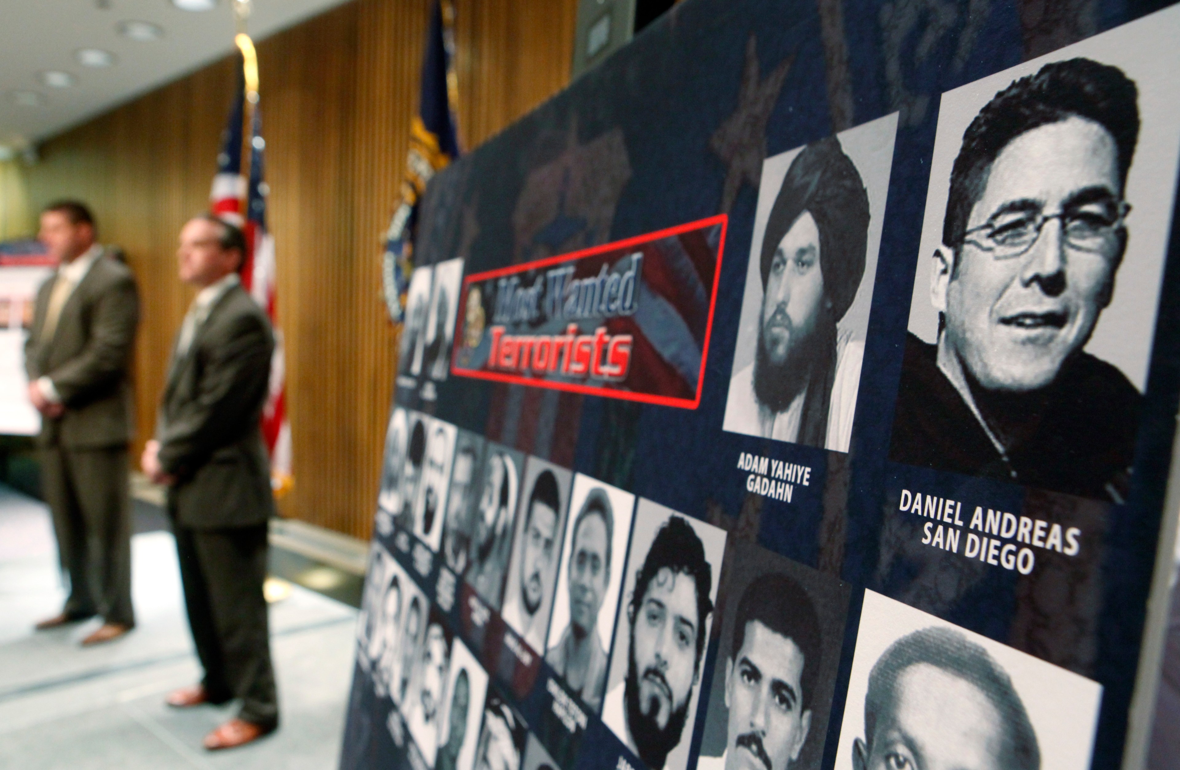 The image shows a "Most Wanted Terrorists" board with headshots and names. Two men stand beside it in suits. American flags are displayed in the background.