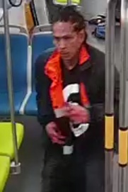 A person with braided hair is on public transport, wearing a black and orange jacket over a shirt with a large white number, holding an object in their hands.