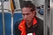 A person with braided hair is on public transport, wearing a black and orange jacket over a shirt with a large white number, holding an object in their hands.