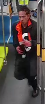 A person with braided hair is on public transport, wearing a black and orange jacket over a shirt with a large white number, holding an object in their hands.