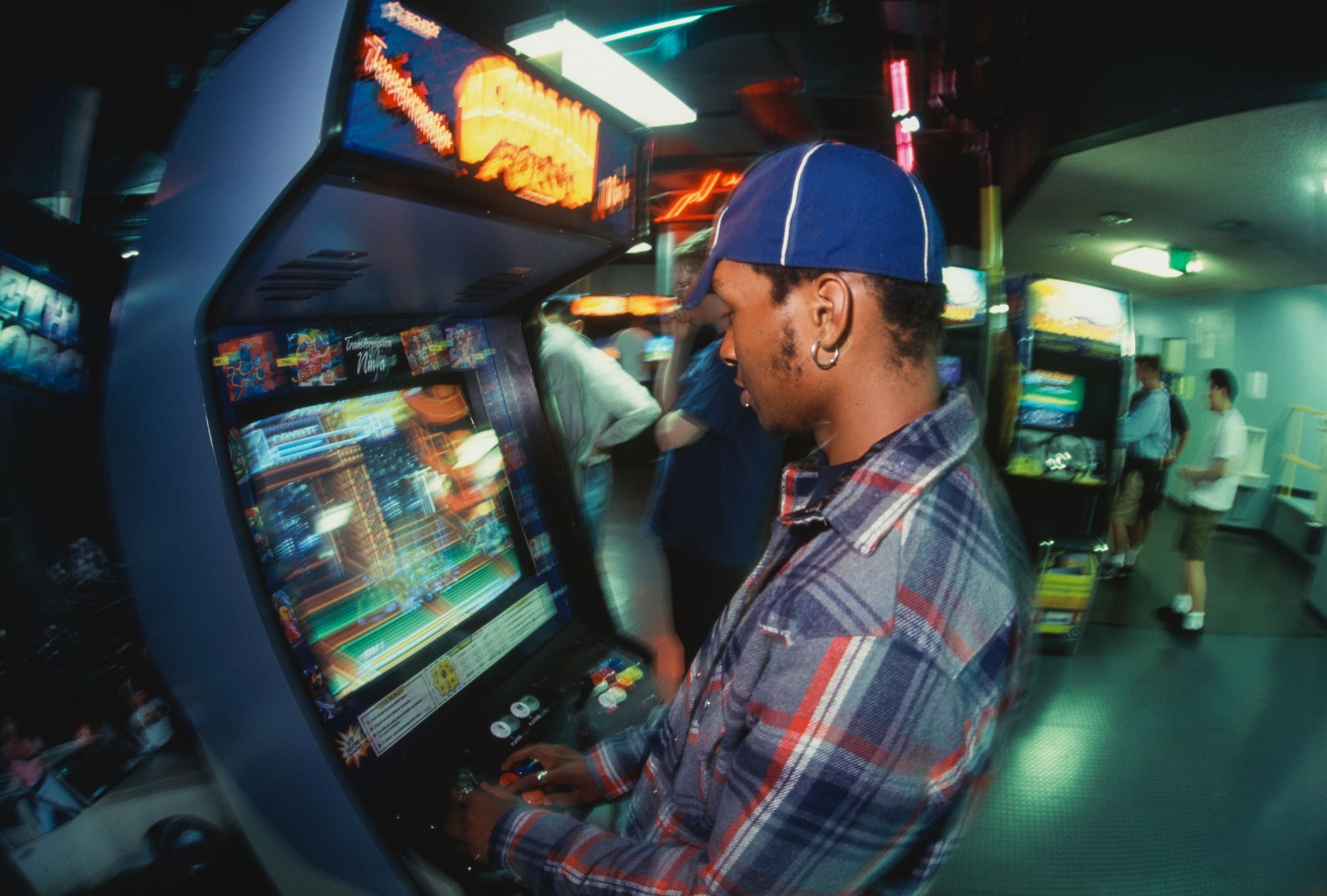 A person in a plaid shirt and blue cap is playing a retro arcade game. The scene is in a dimly lit arcade with others in the background.