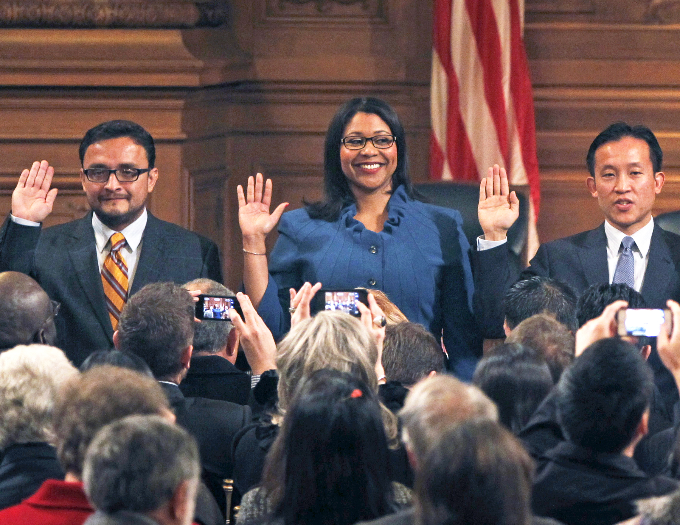 Three individuals stand in front of an audience with their right hands raised, appearing to take an oath. A U.S. flag is visible in the background.