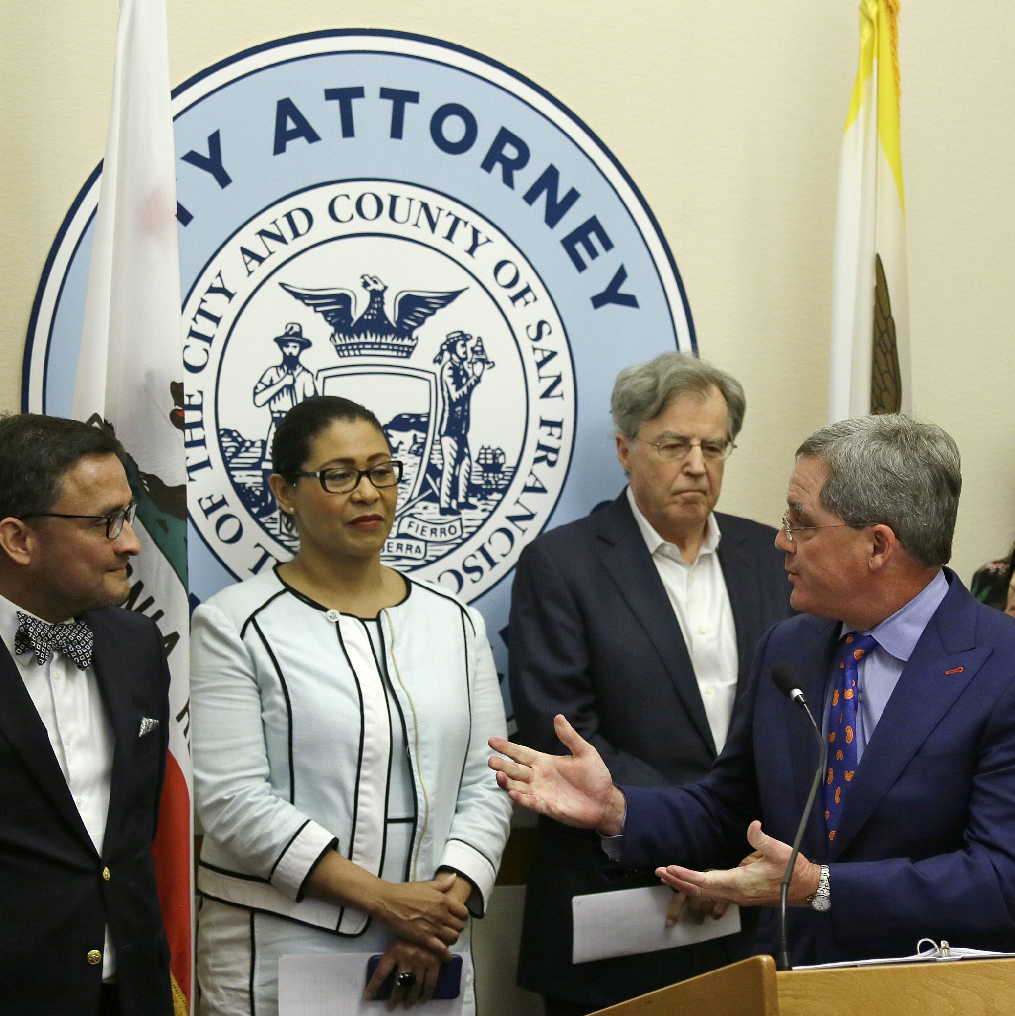 Several individuals in formal attire stand in front of a San Francisco City Attorney seal, with U.S. and California flags visible behind them.