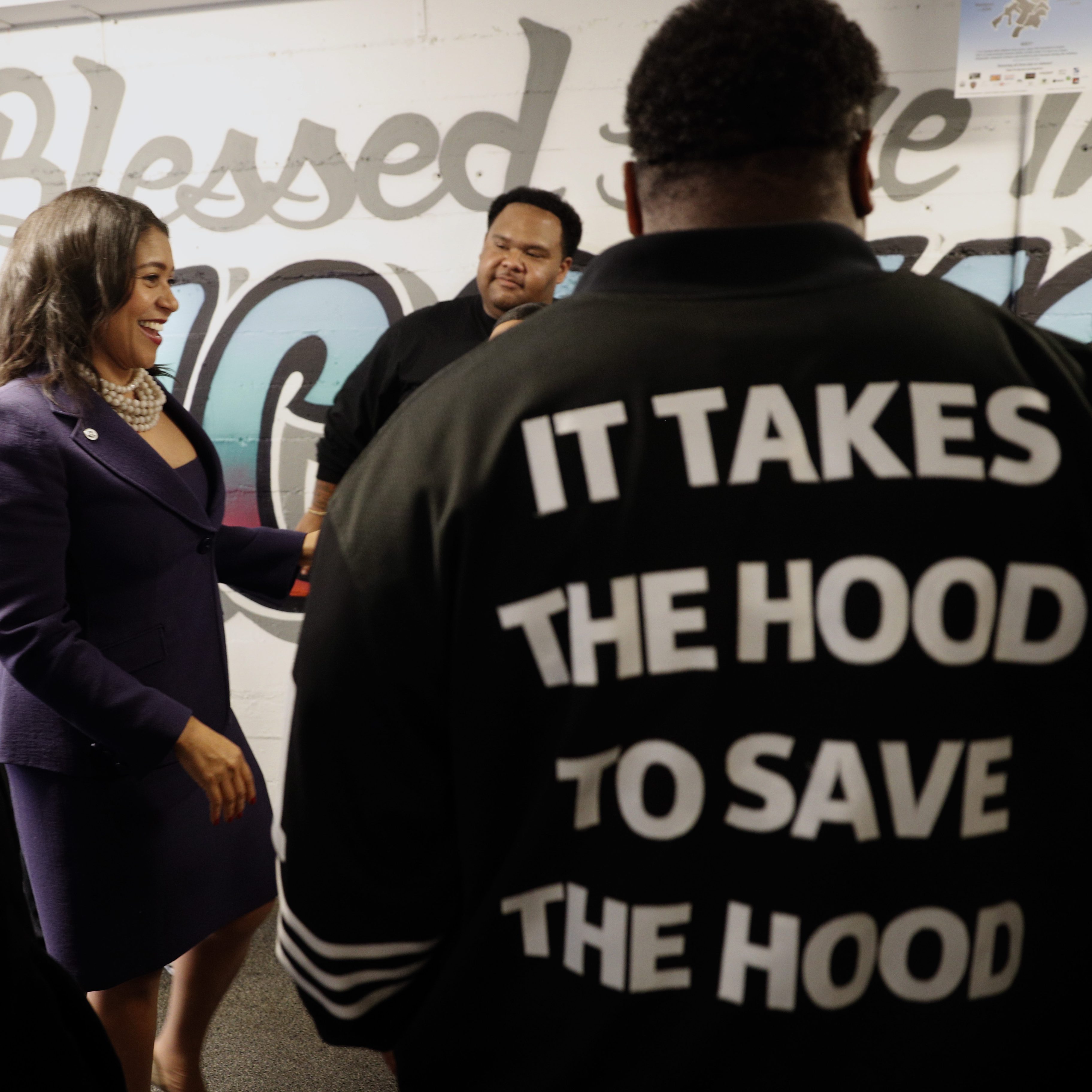 A group of people is gathered in a room with colorful graffiti on the wall. One person wears a jacket that says &quot;It takes the hood to save the hood&quot; on the back.