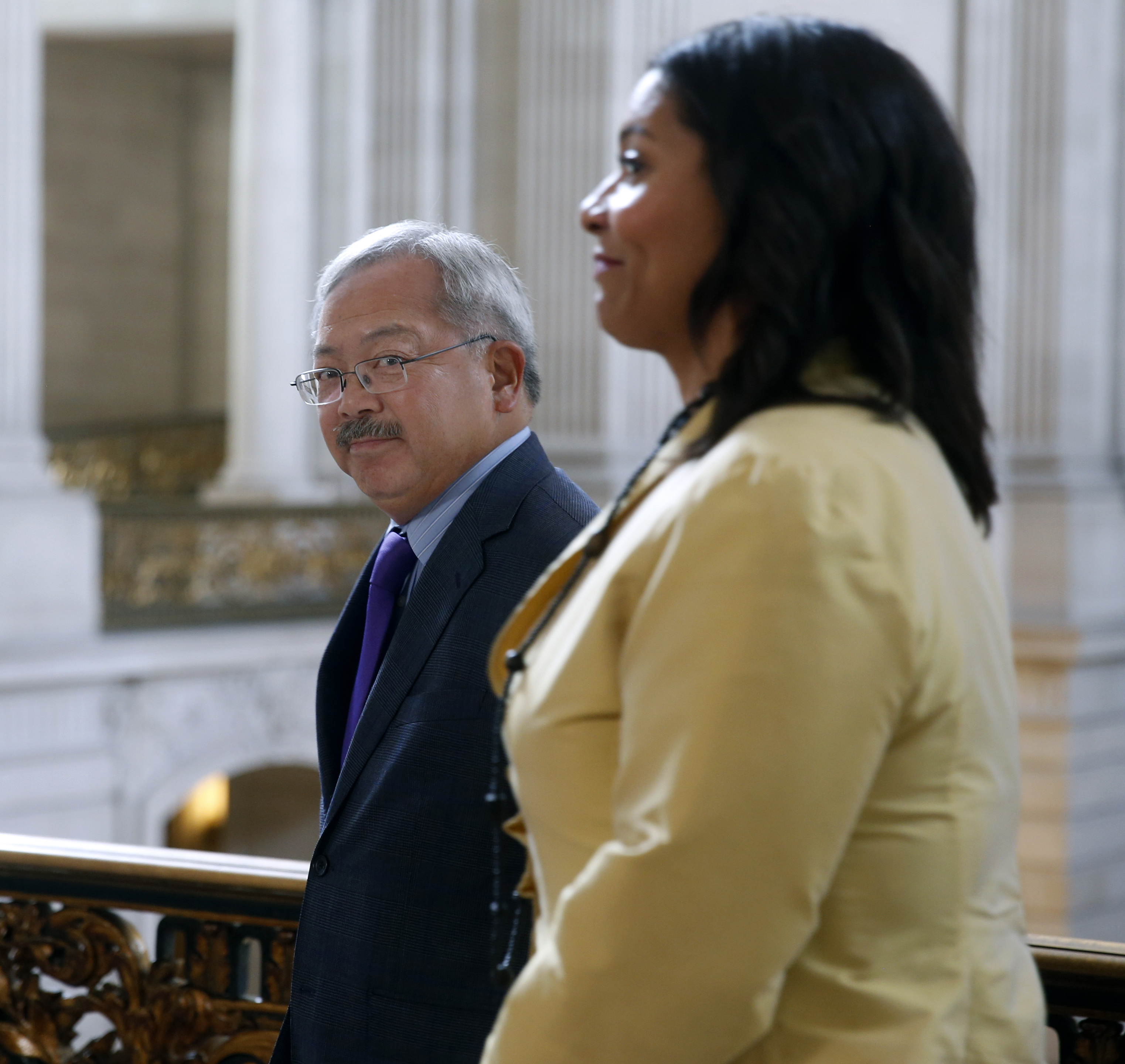 Two people stand indoors, the man in a suit with a purple tie. The woman, in a yellow jacket, is slightly closer. The background features ornate architecture.