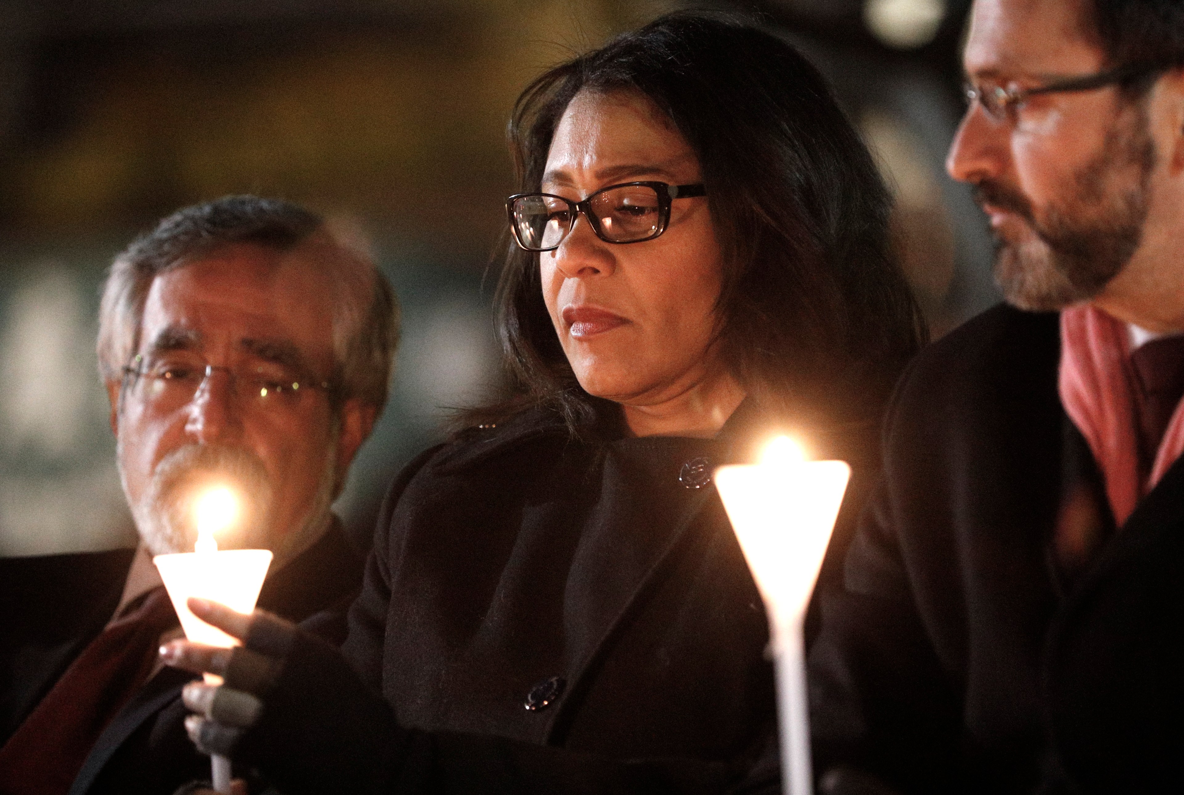 Three people sit closely together, each holding a lit candle. They appear solemn, and the glow from the candles illuminates their faces.