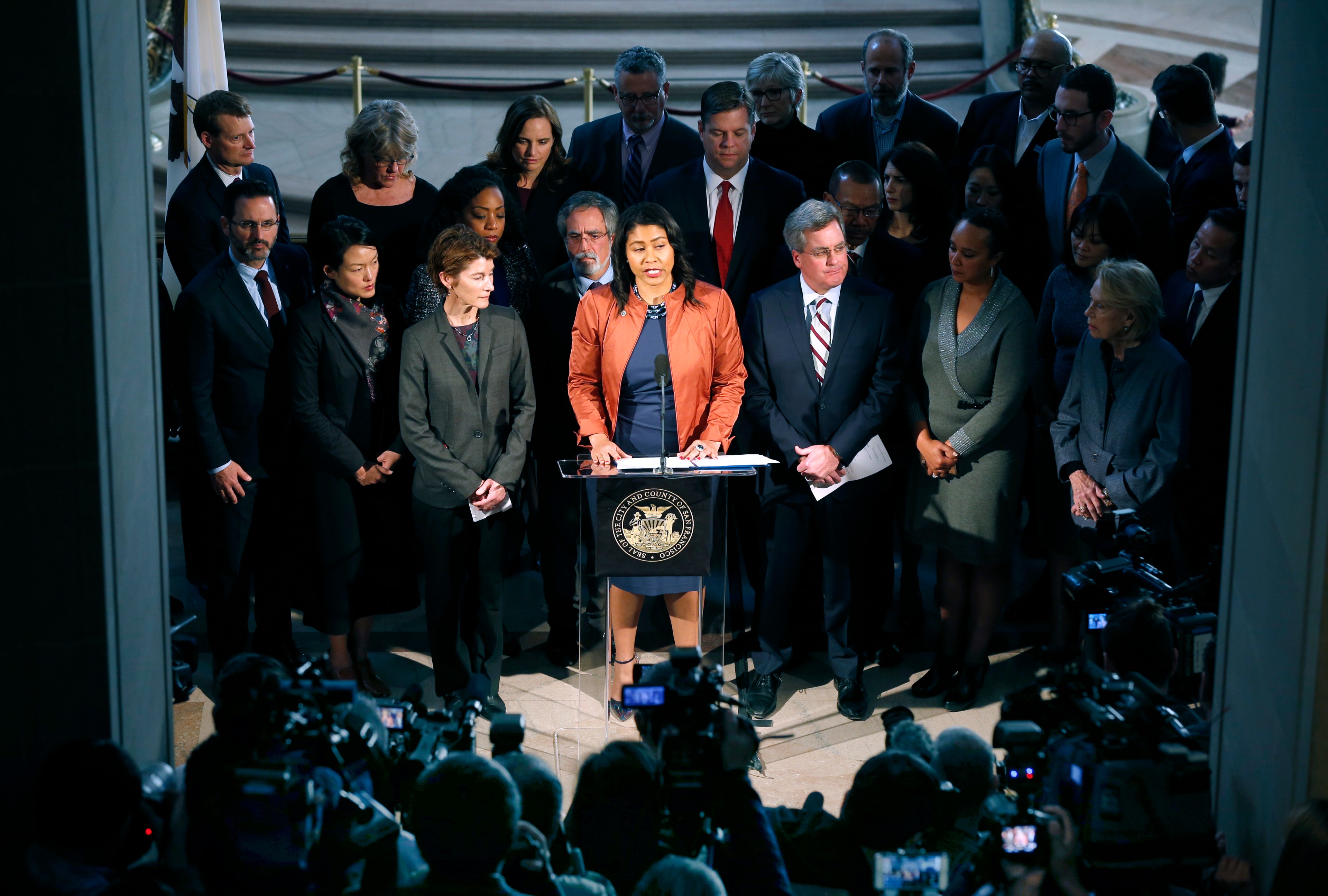 A woman speaks at a podium surrounded by a large group of people. Cameras capture the event in a formal setting with a government emblem on the podium.