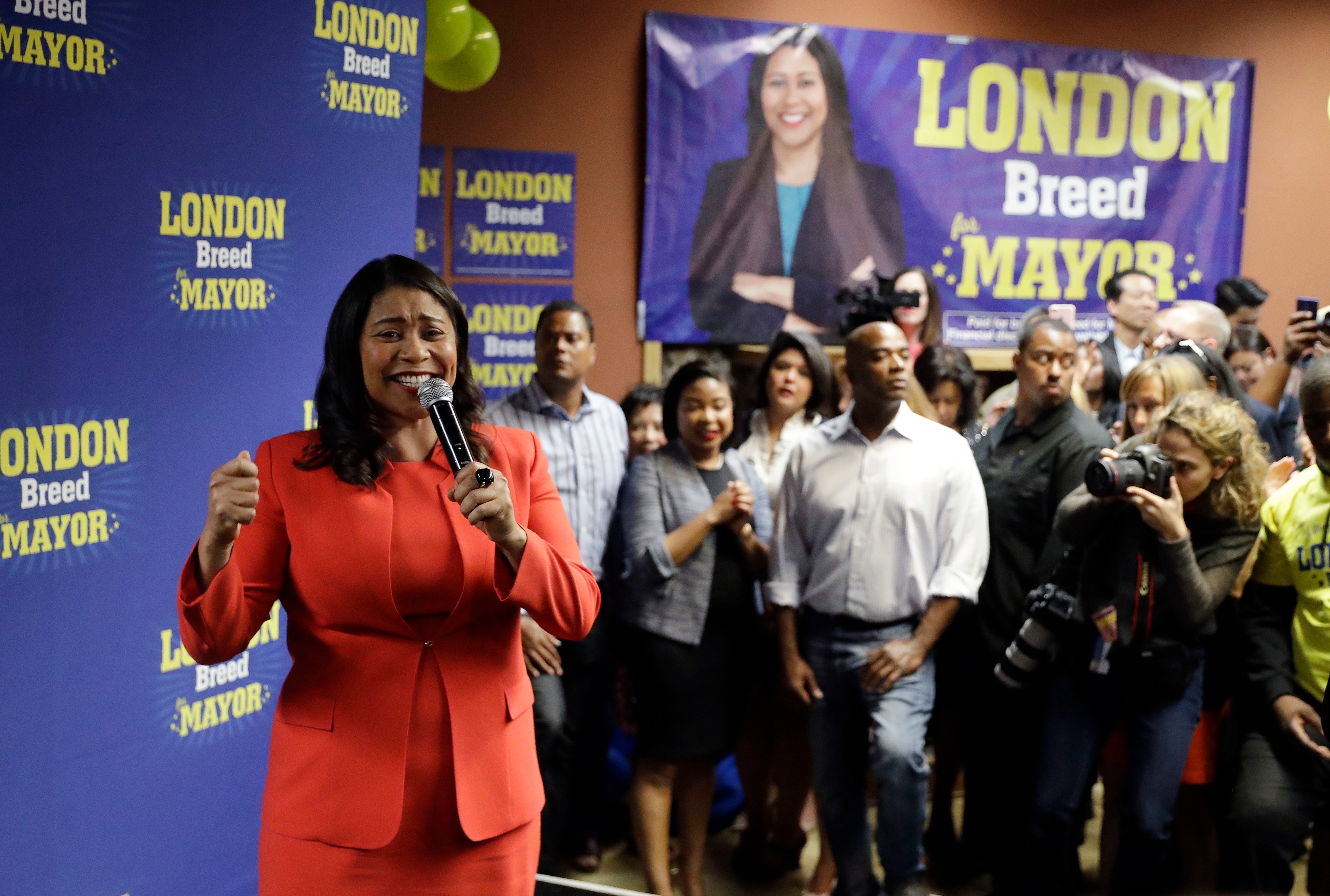 A woman in a red outfit is speaking into a microphone at a campaign event with a supportive crowd and a large poster behind her.