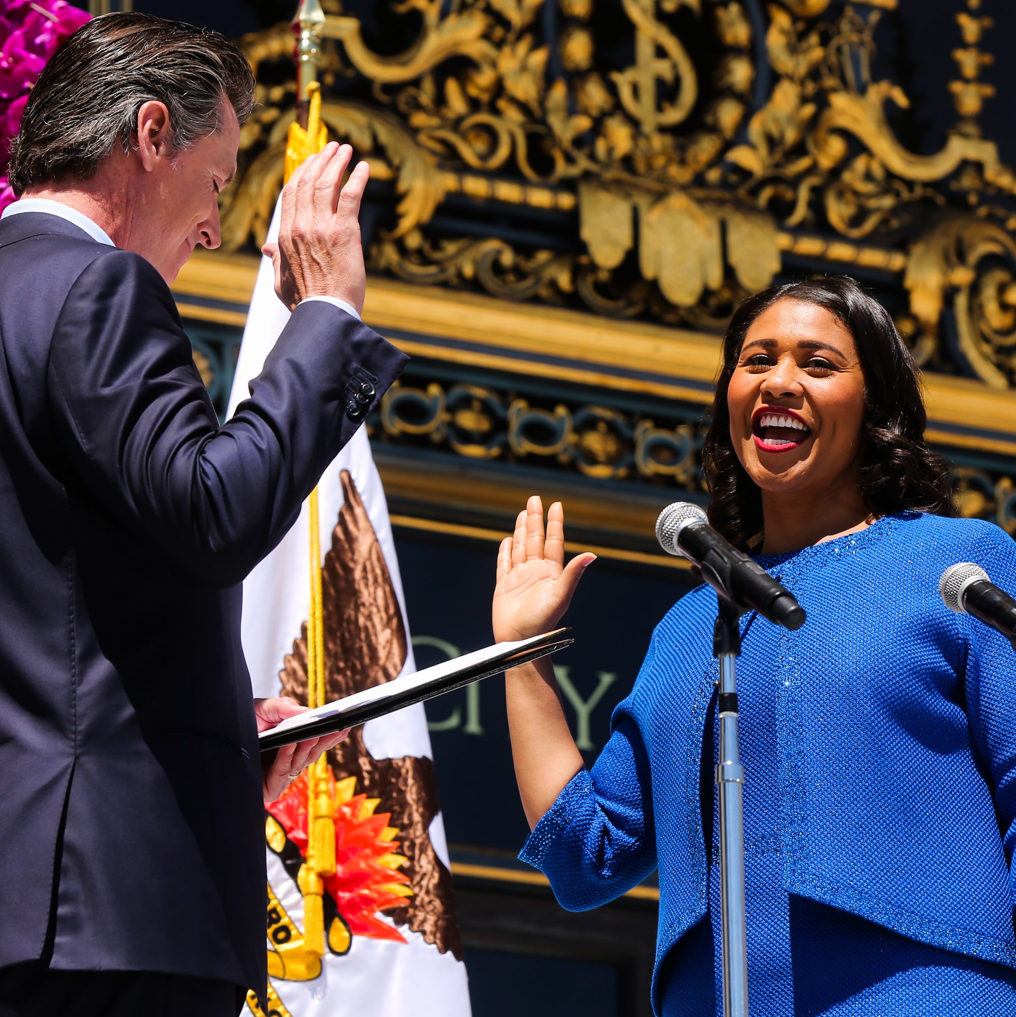 Two people are in front of elaborate gold decorations, raising their hands in a ceremony. One is holding a document, and there are microphones and flags nearby.