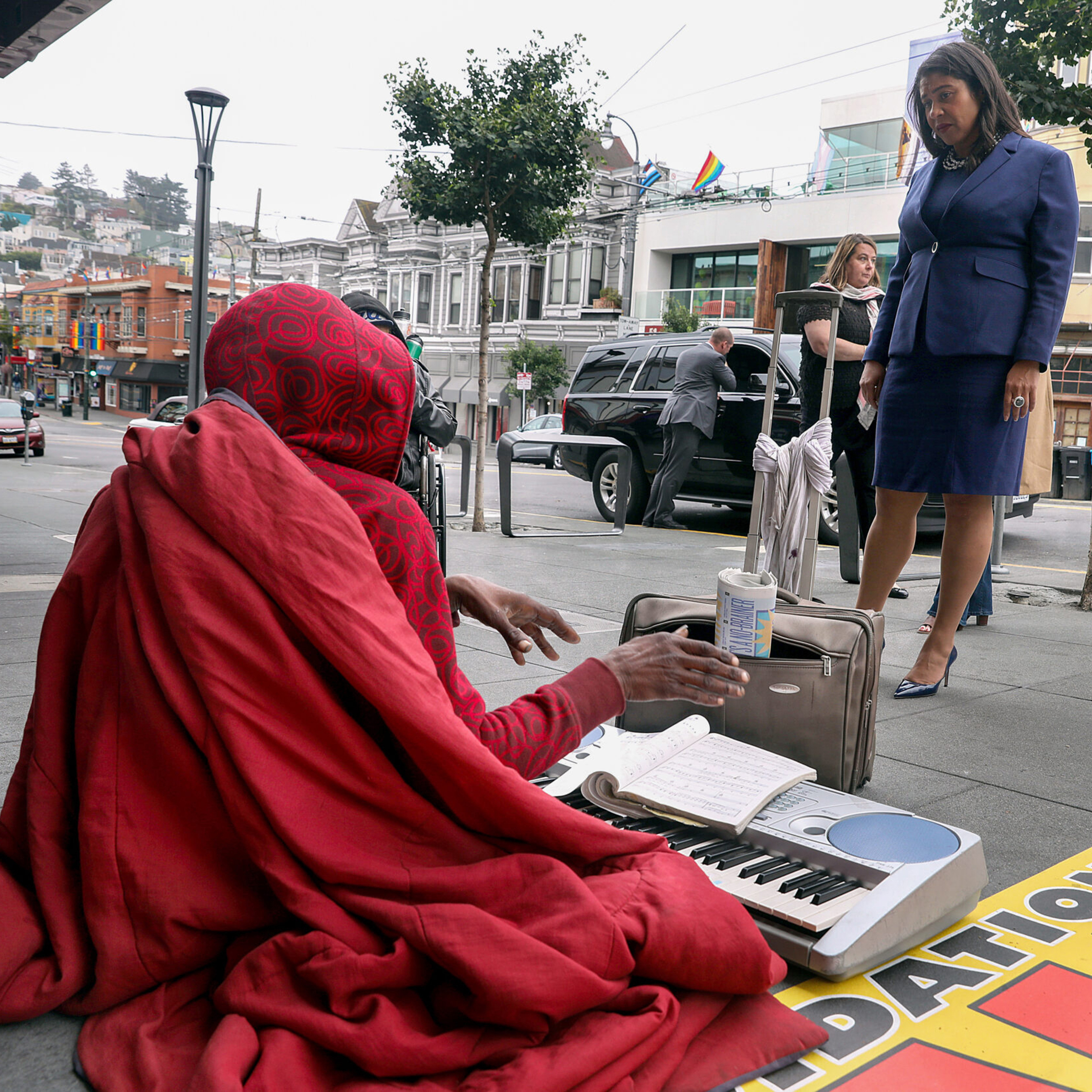 A person in a red hoodie sits on the sidewalk with a keyboard and notebook, engaging with a woman in a blue suit, as people and vehicles pass by.