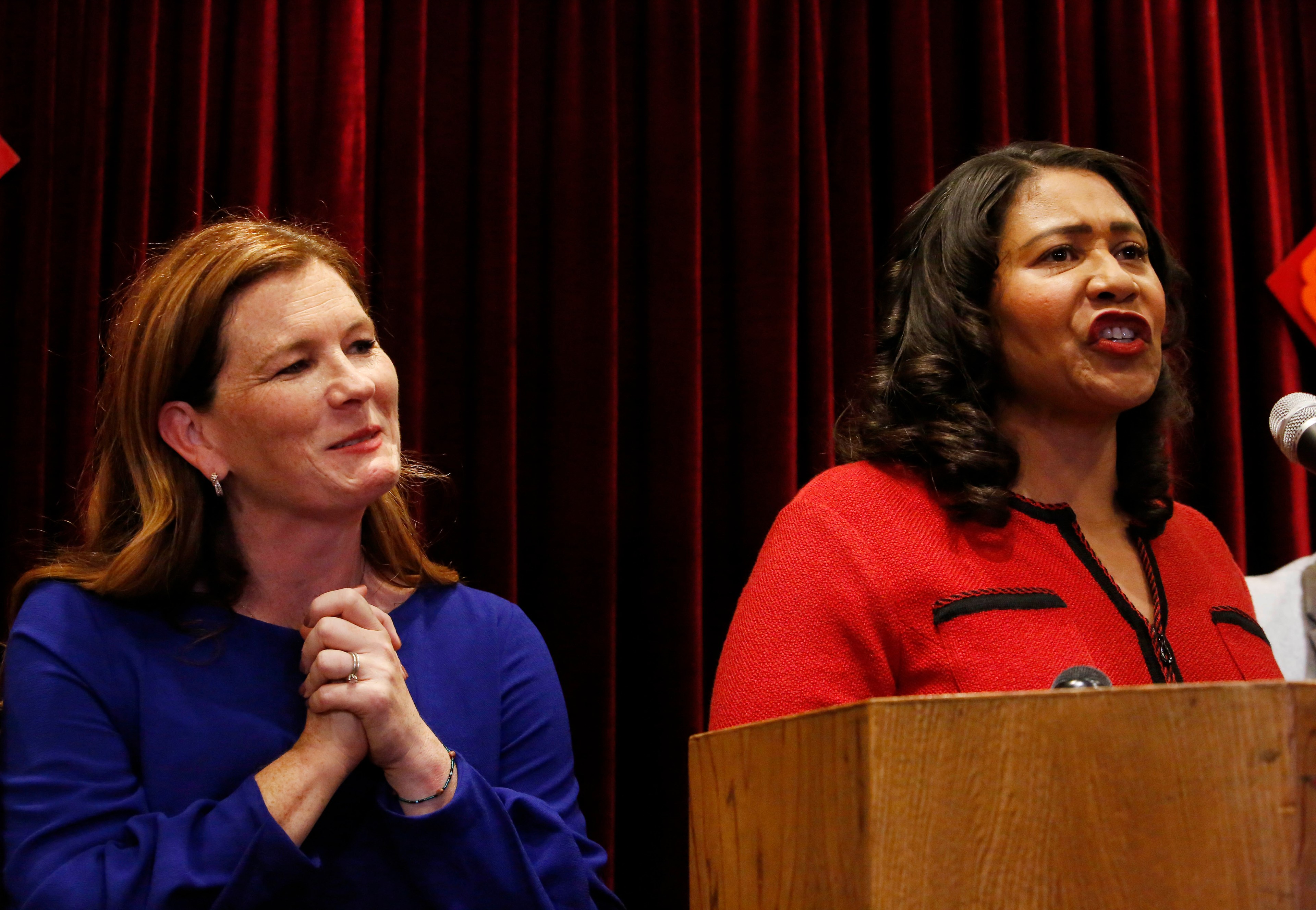 Two women are in front of a red curtain. One is speaking at a podium, wearing a red outfit, and the other stands next to her in blue, smiling and clasping her hands.