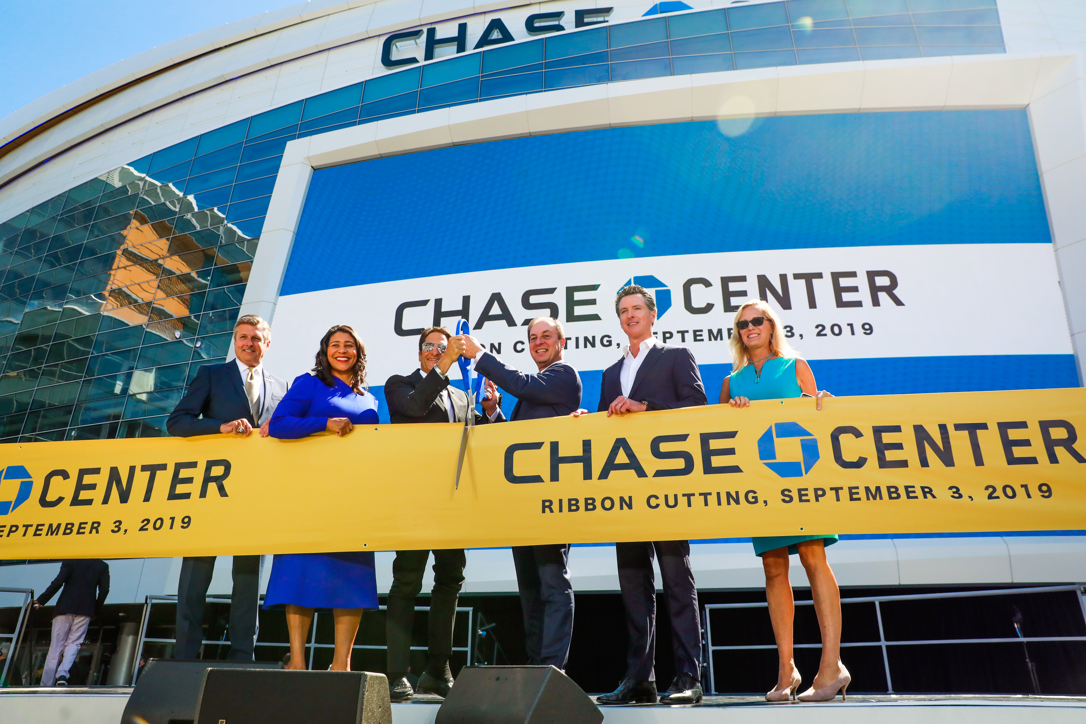 A group of people stand smiling in front of Chase Center, holding a large yellow ribbon with &quot;Ribbon Cutting, September 3, 2019&quot; written on it.