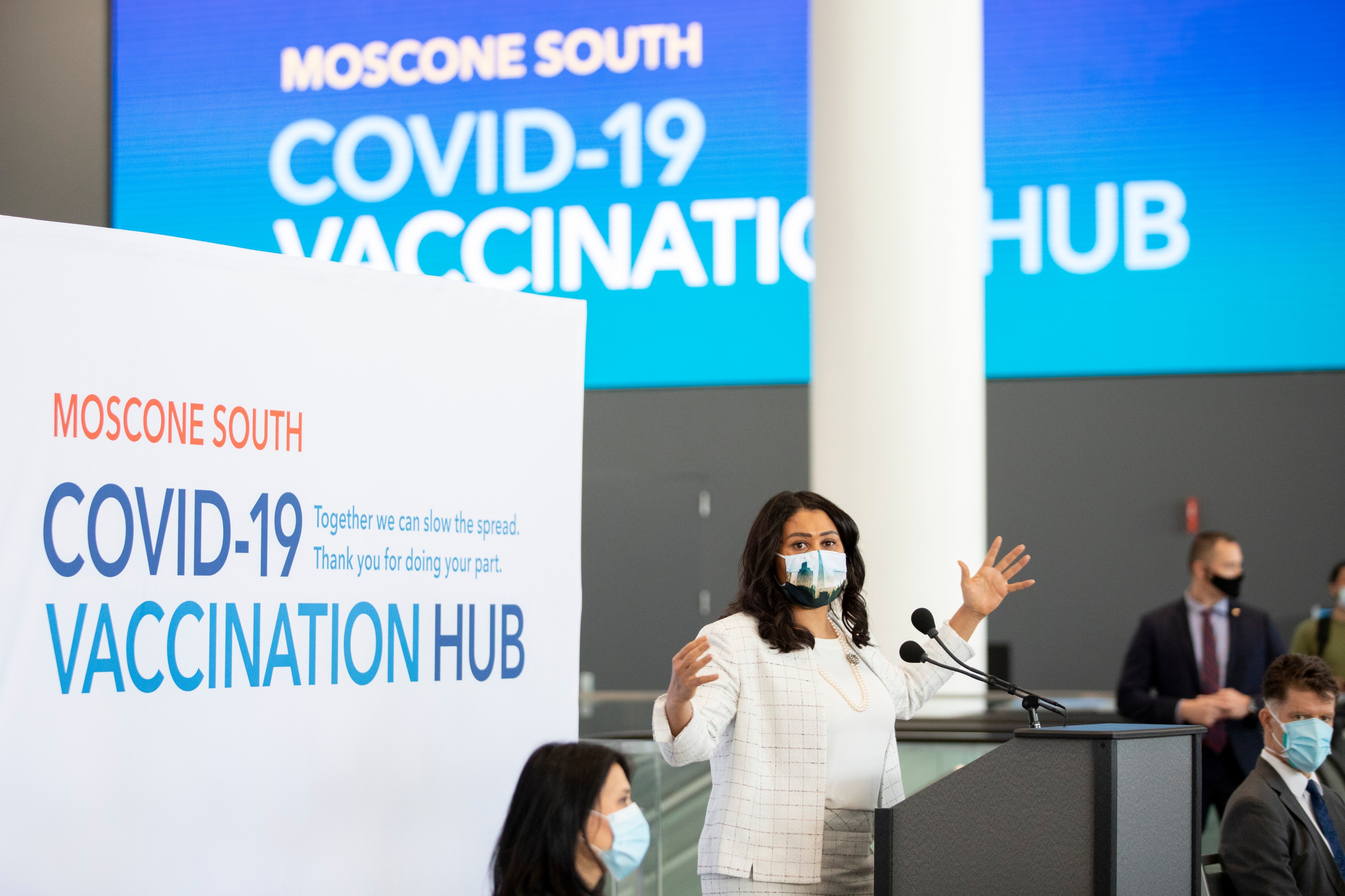 A woman speaks at a podium wearing a mask in a COVID-19 vaccination hub, with a large sign and backdrop displaying &quot;MOSCONE SOUTH COVID-19 VACCINATION HUB.&quot;