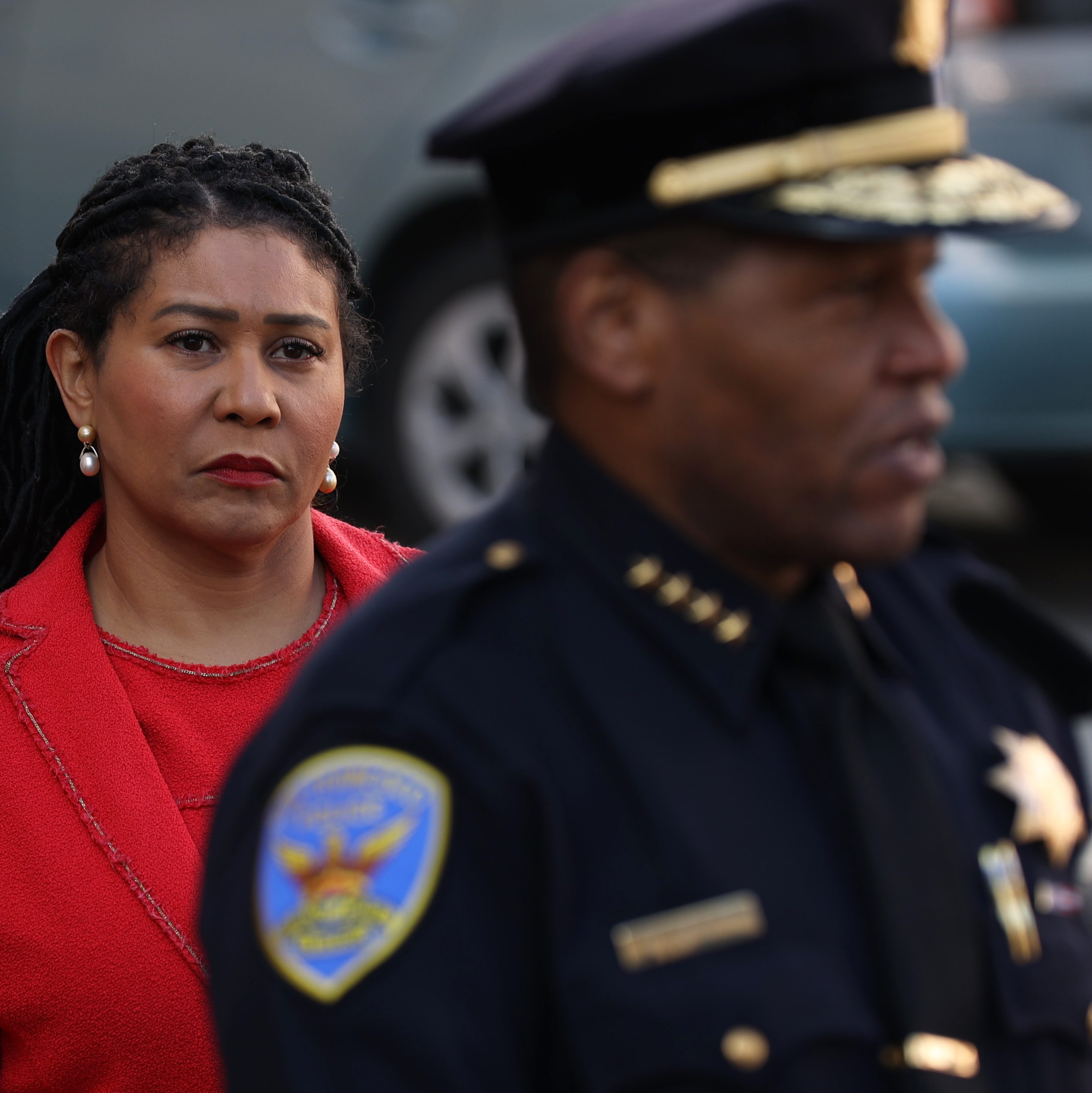 A woman in a red coat with pearl earrings looks serious as she stands outside. In the foreground, a uniformed police officer is partially visible.