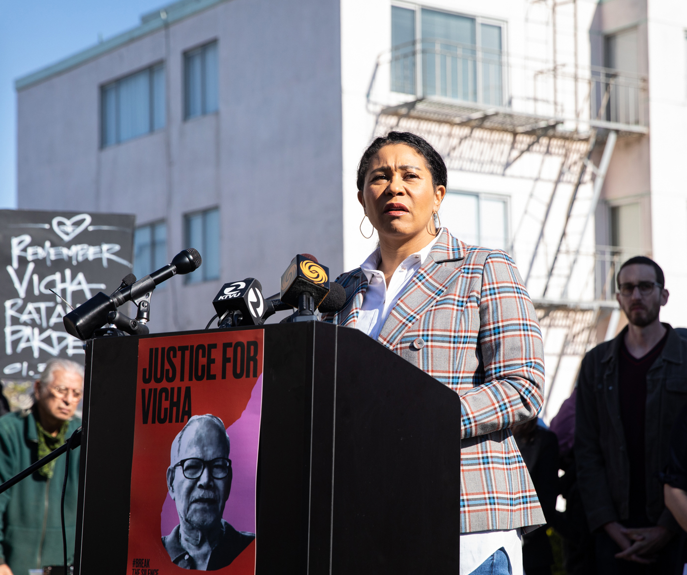 A woman speaks at a podium with microphones, featuring a poster that reads &quot;Justice for Vicha.&quot; A &quot;Remember Vicha Ratanapakdee&quot; sign is also visible.