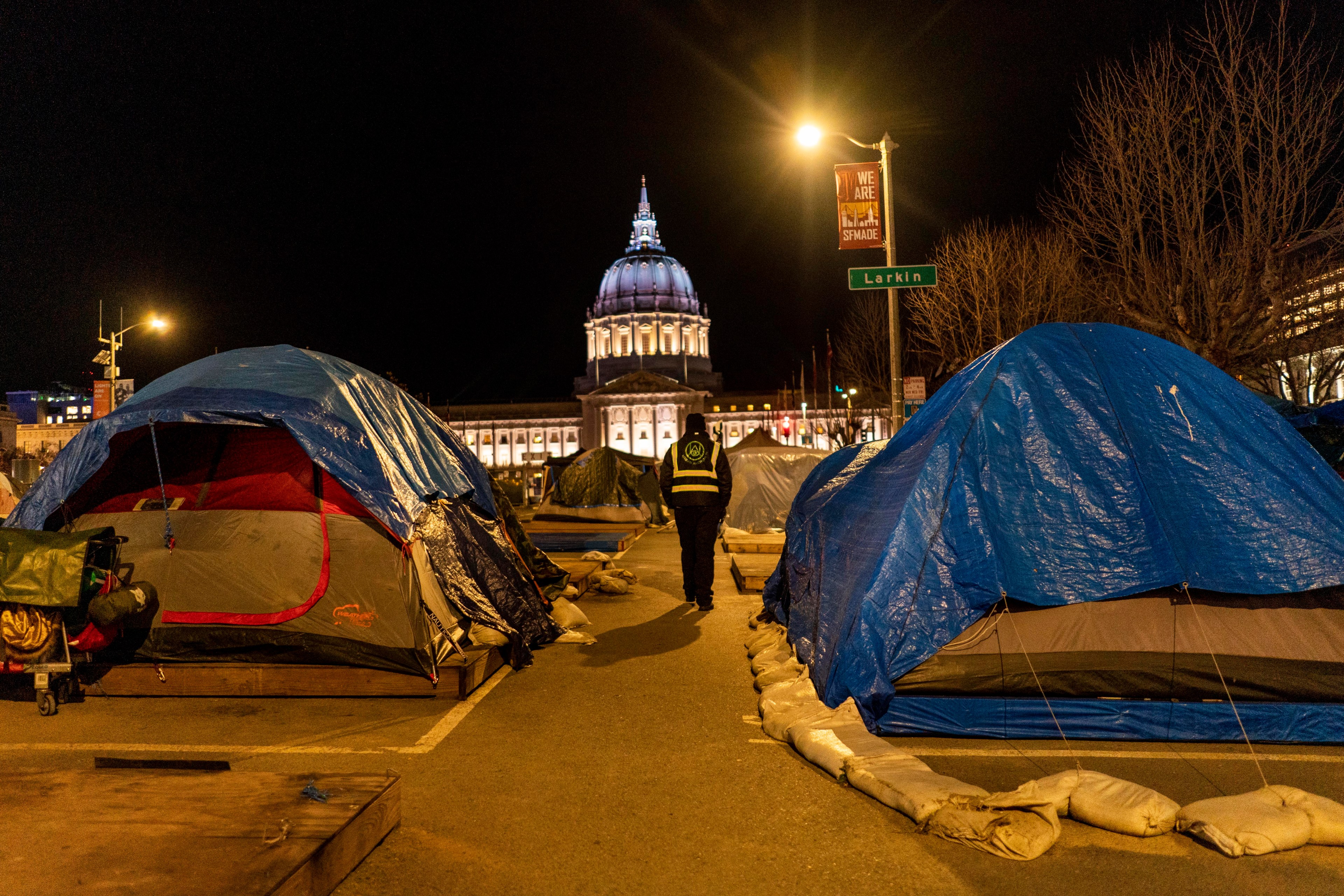 A row of tents covered with blue tarps is set up on a street at night. A person in a vest walks by, with a domed, illuminated building in the background.