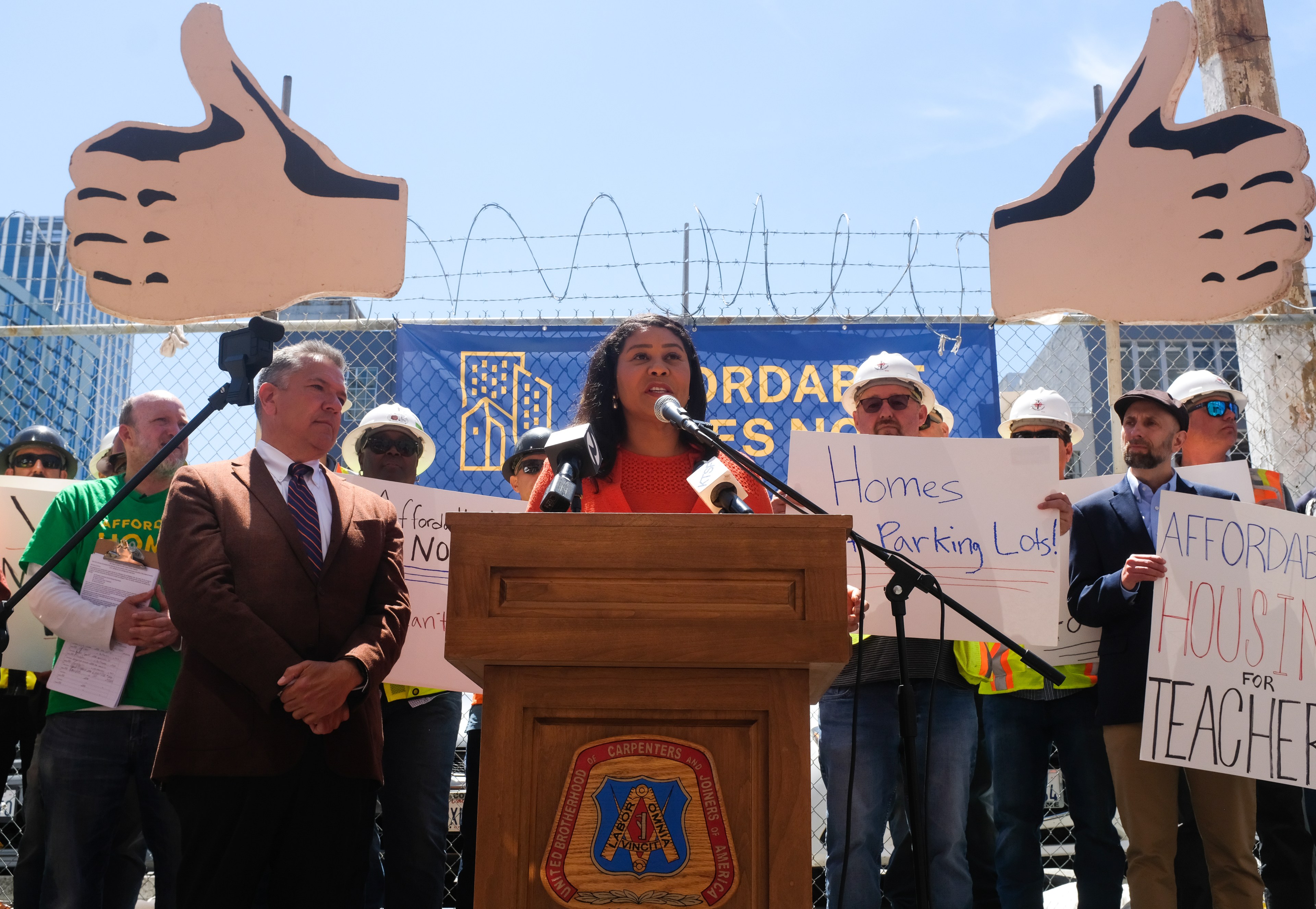 A woman speaks at a podium surrounded by people holding signs about affordable housing. Two large foam thumbs-up signs are in the background.