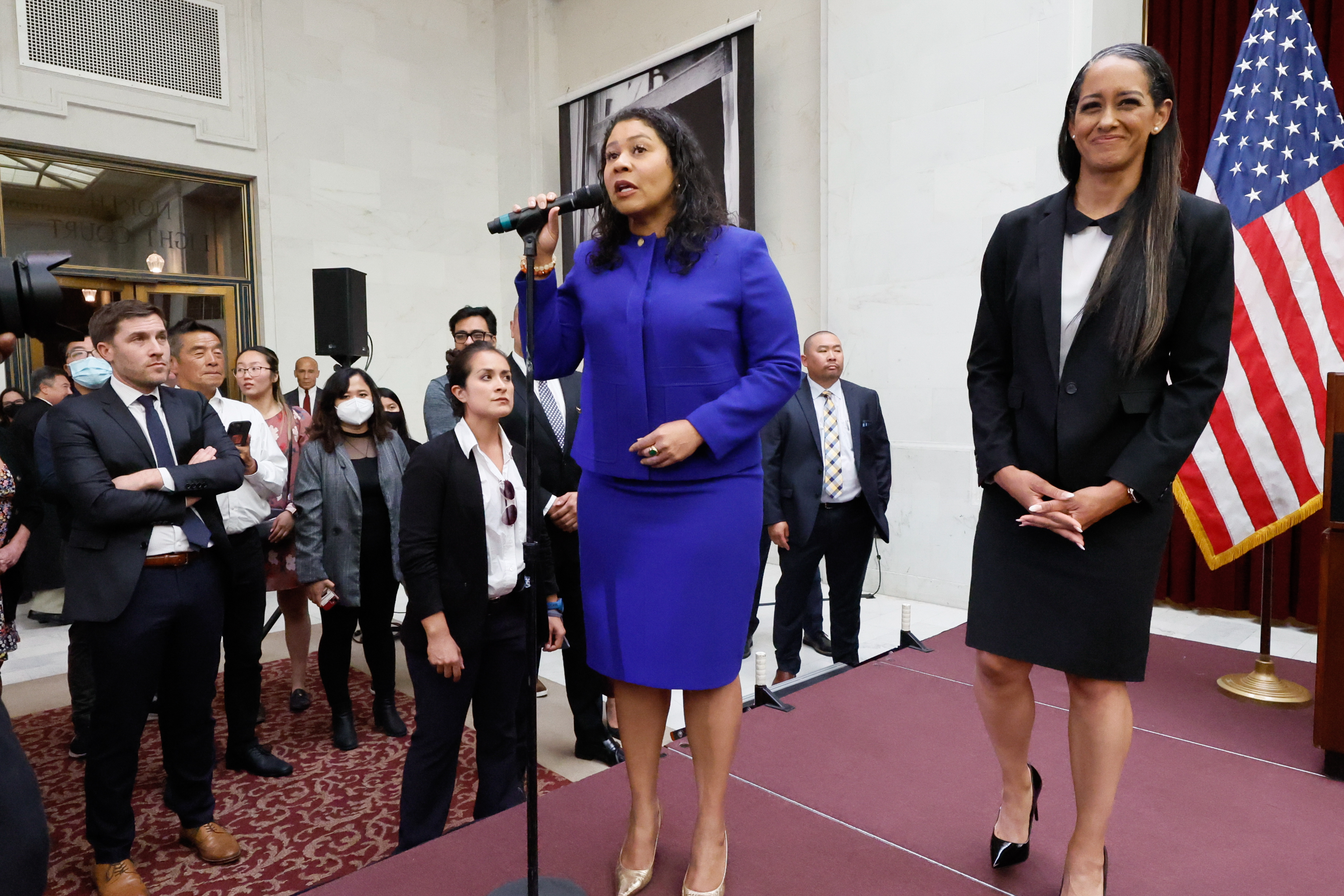 A woman in a blue outfit speaks into a microphone on a stage, surrounded by a crowd. Another woman in a black suit stands beside her, with a U.S. flag in the background.