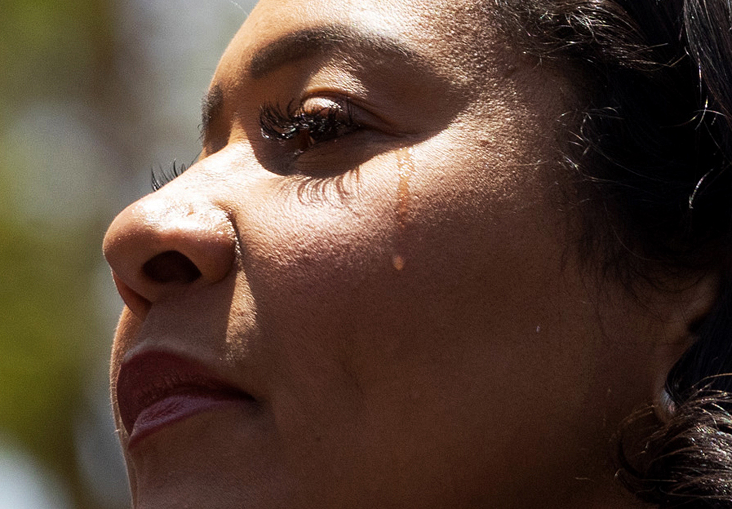 The image shows a close-up of a person’s face in profile with a tear rolling down their cheek, set against an outdoor background.