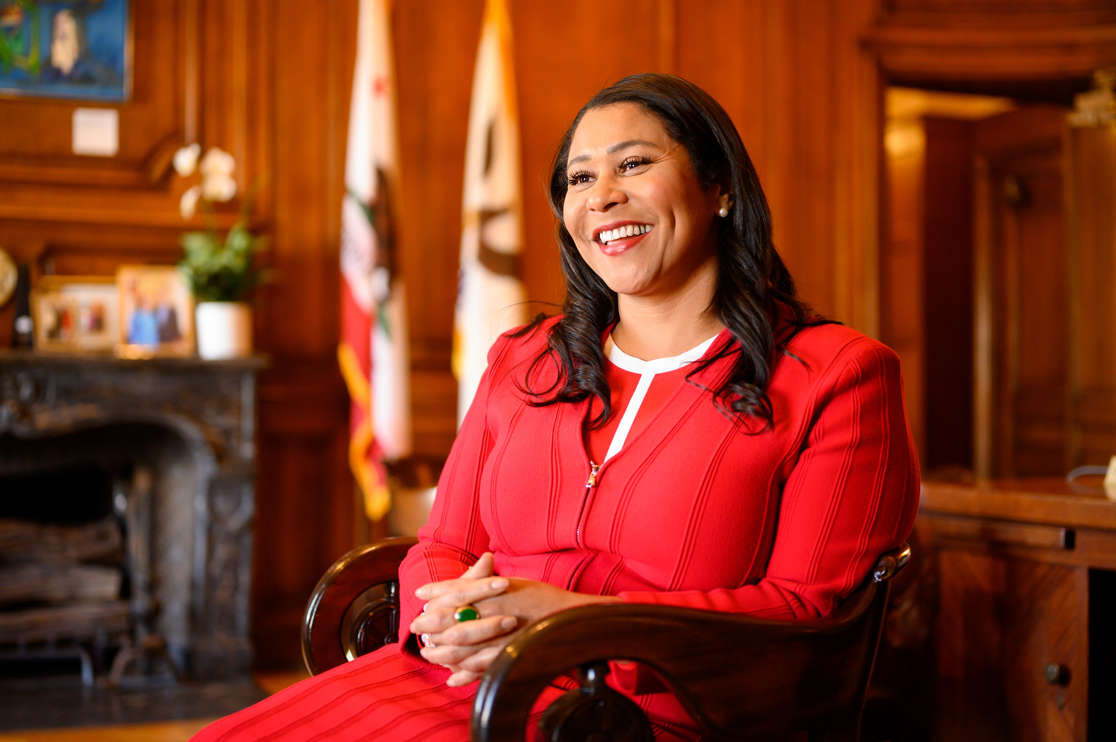 A person in a red suit smiles warmly, seated in a wood-paneled room with flags and framed photos in the background, creating a formal yet inviting atmosphere.