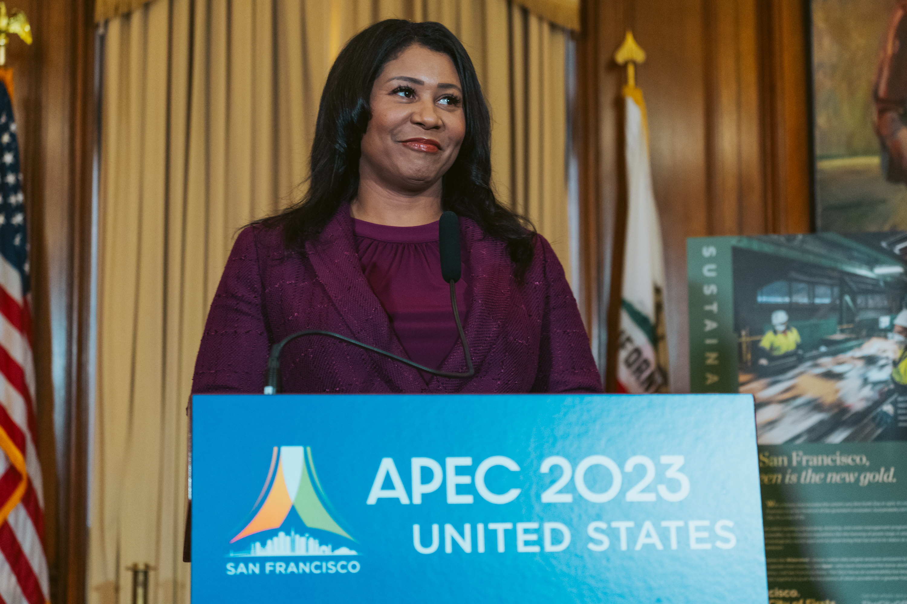 A woman stands at a podium labeled &quot;APEC 2023 United States&quot; with a San Francisco logo. The setting includes flags and a poster in the background.