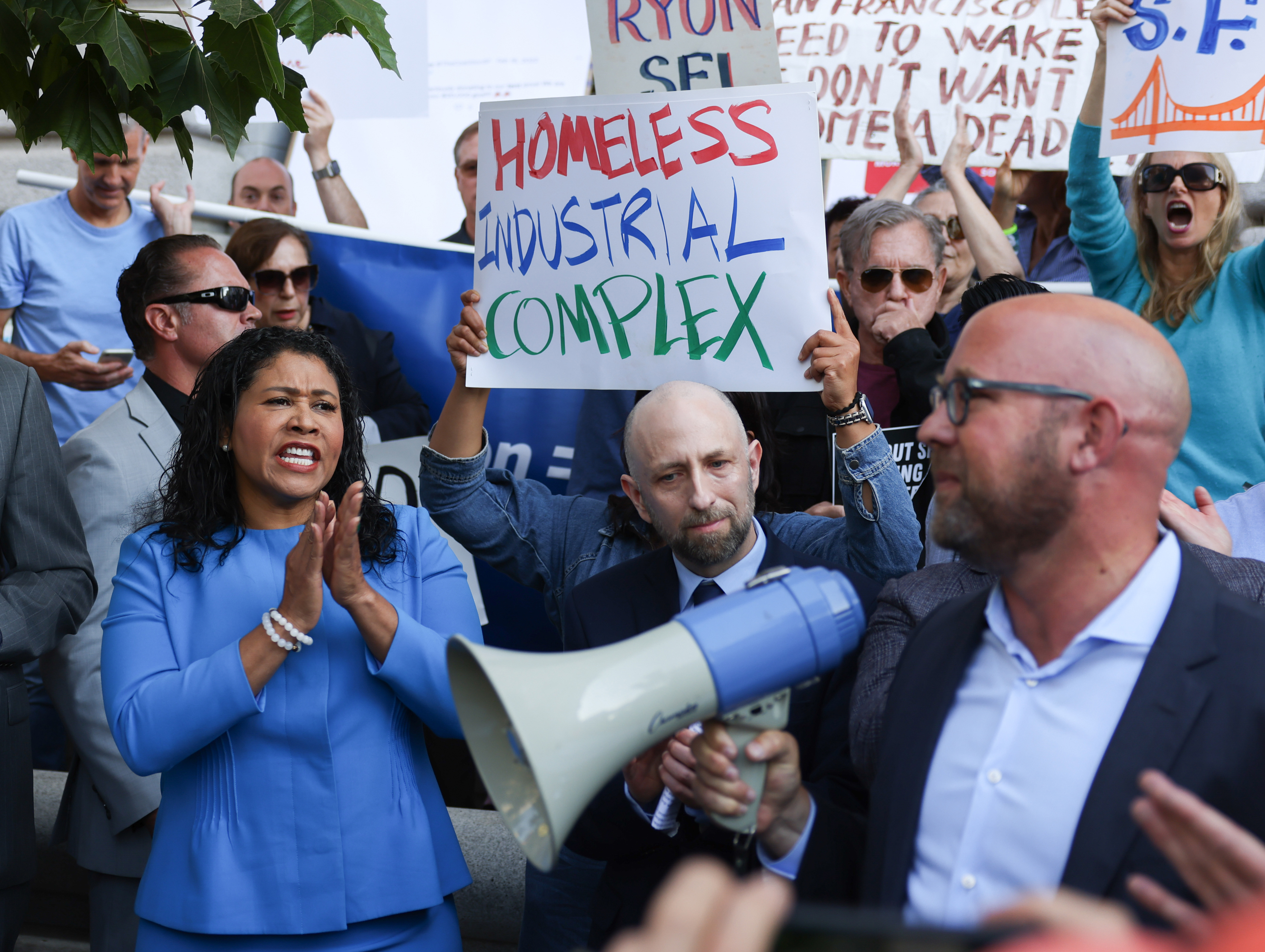 A group of people, including a woman in a blue suit speaking and clapping, gather with protest signs about homelessness. One sign reads &quot;Homeless Industrial Complex.&quot;