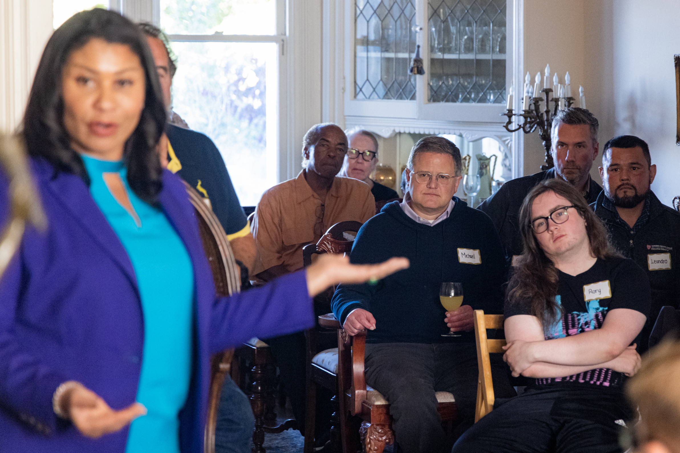 A woman in the foreground gestures while a group of people, including two with visible nametags, attentively sit behind her in a room with a window and cabinet.