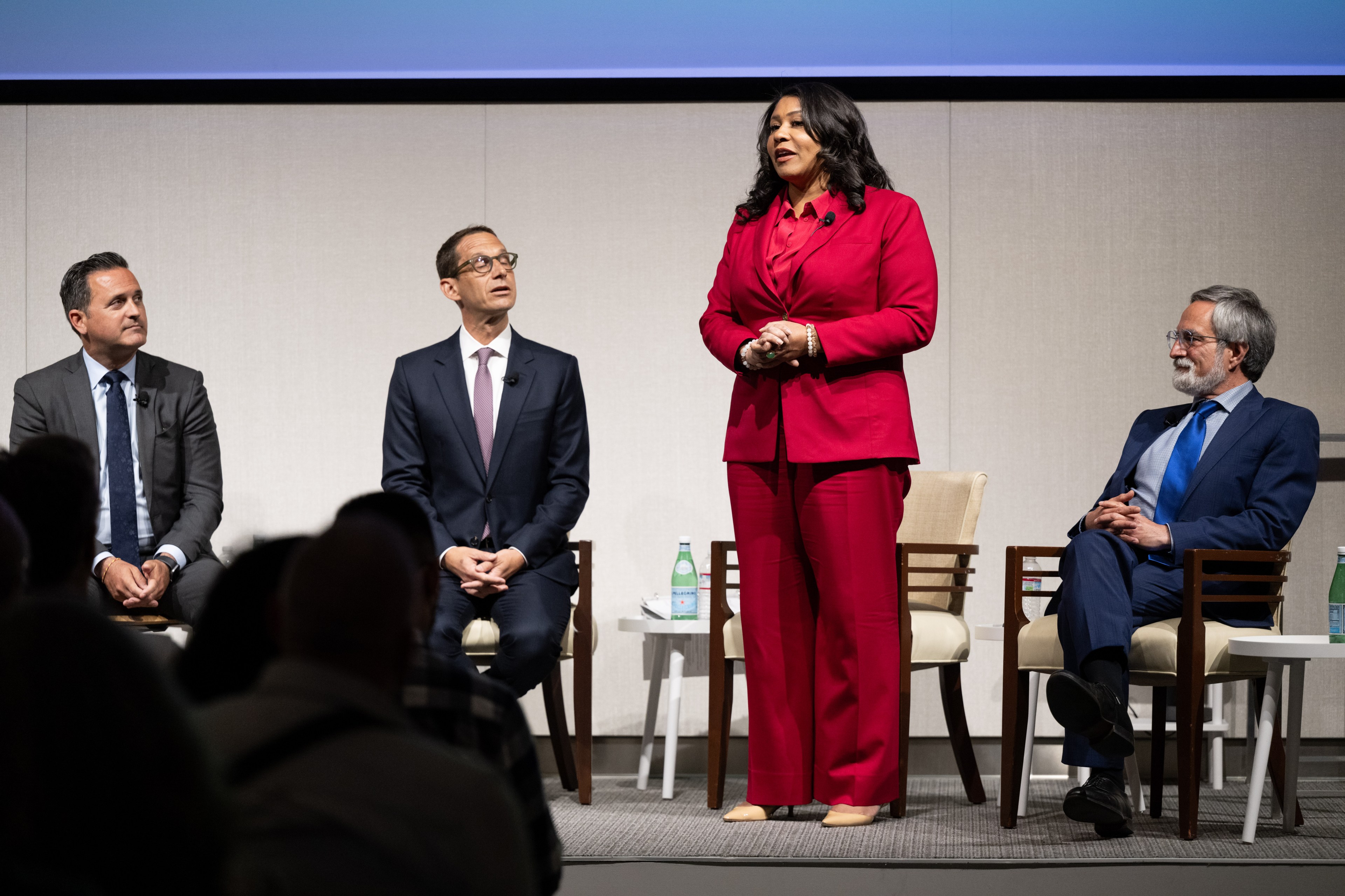 Four people in professional attire are on a stage. Three are seated in chairs, while a woman in a red suit stands speaking. Bottles of water are on tables nearby.