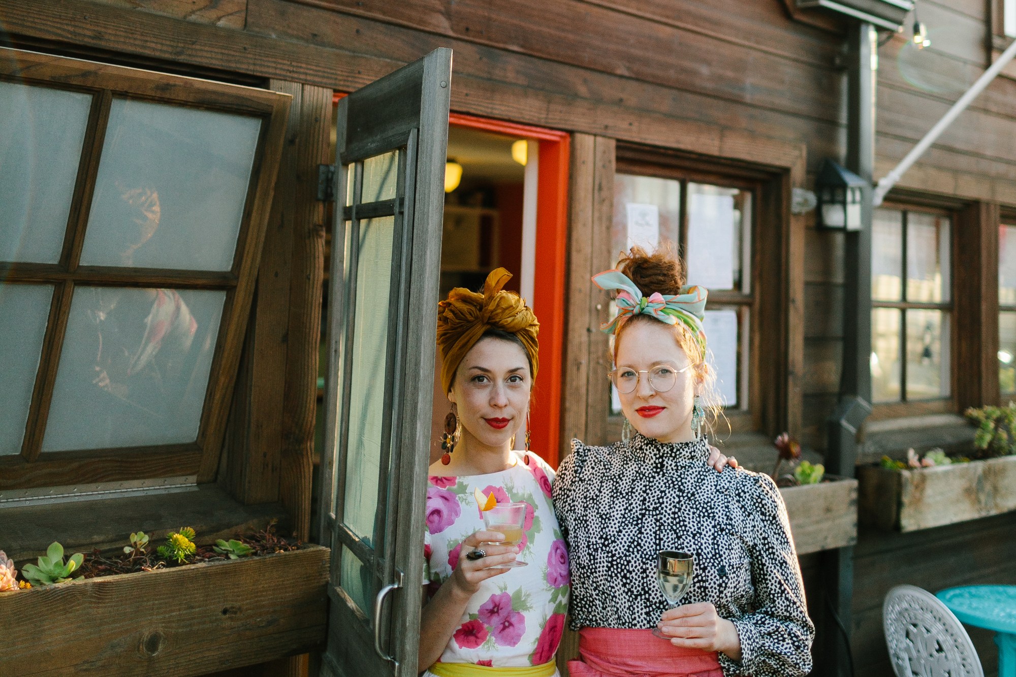 Two people stand smiling outside a rustic cafe, holding drinks. The sign reads "Fog Eater Cafe". They're dressed vibrantly, with string lights above.