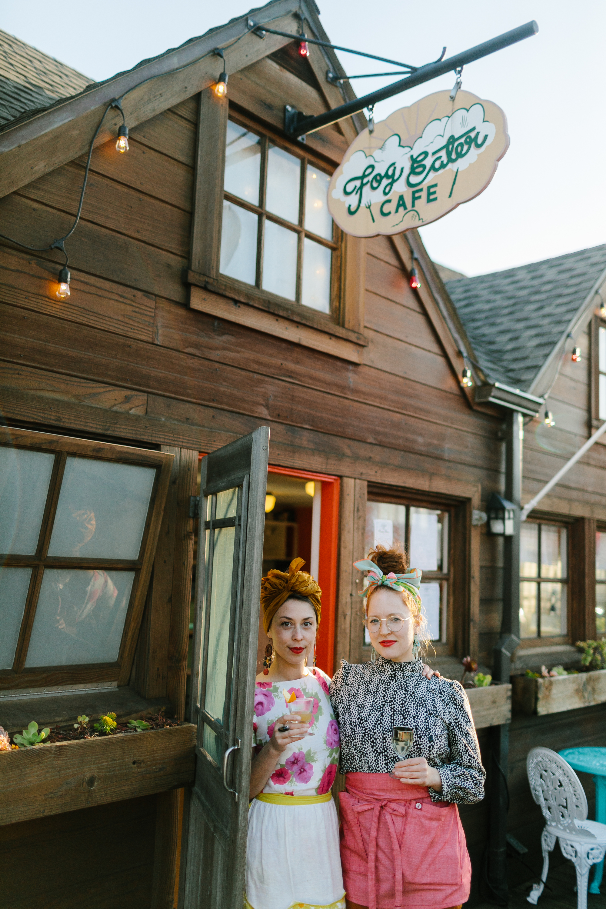 Two people stand smiling outside a rustic cafe, holding drinks. The sign reads "Fog Eater Cafe". They're dressed vibrantly, with string lights above.