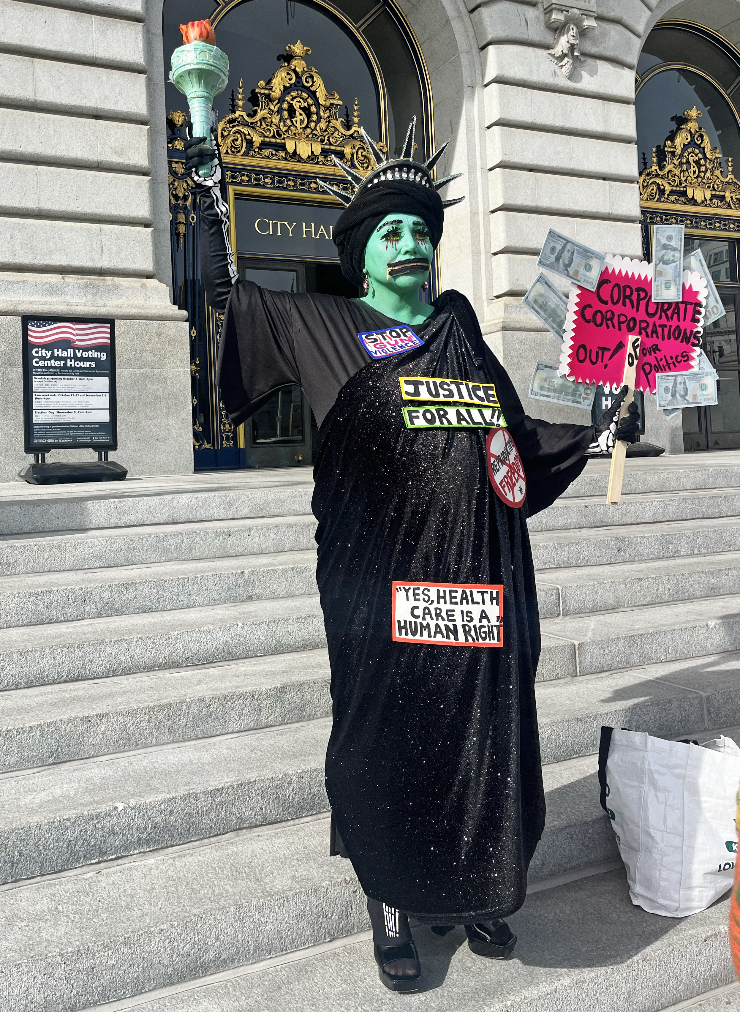 A person dressed as the Statue of Liberty, painted green, holds a sign about corporate influence in politics. They stand on City Hall steps with advocacy stickers.