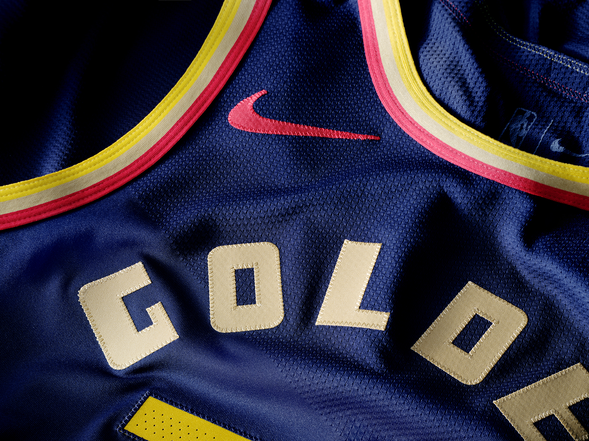 The image shows a close-up of a navy sports jersey with red and yellow trim and stitched letters reading &quot;GOLD.&quot; A red Nike logo is visible near the neckline.