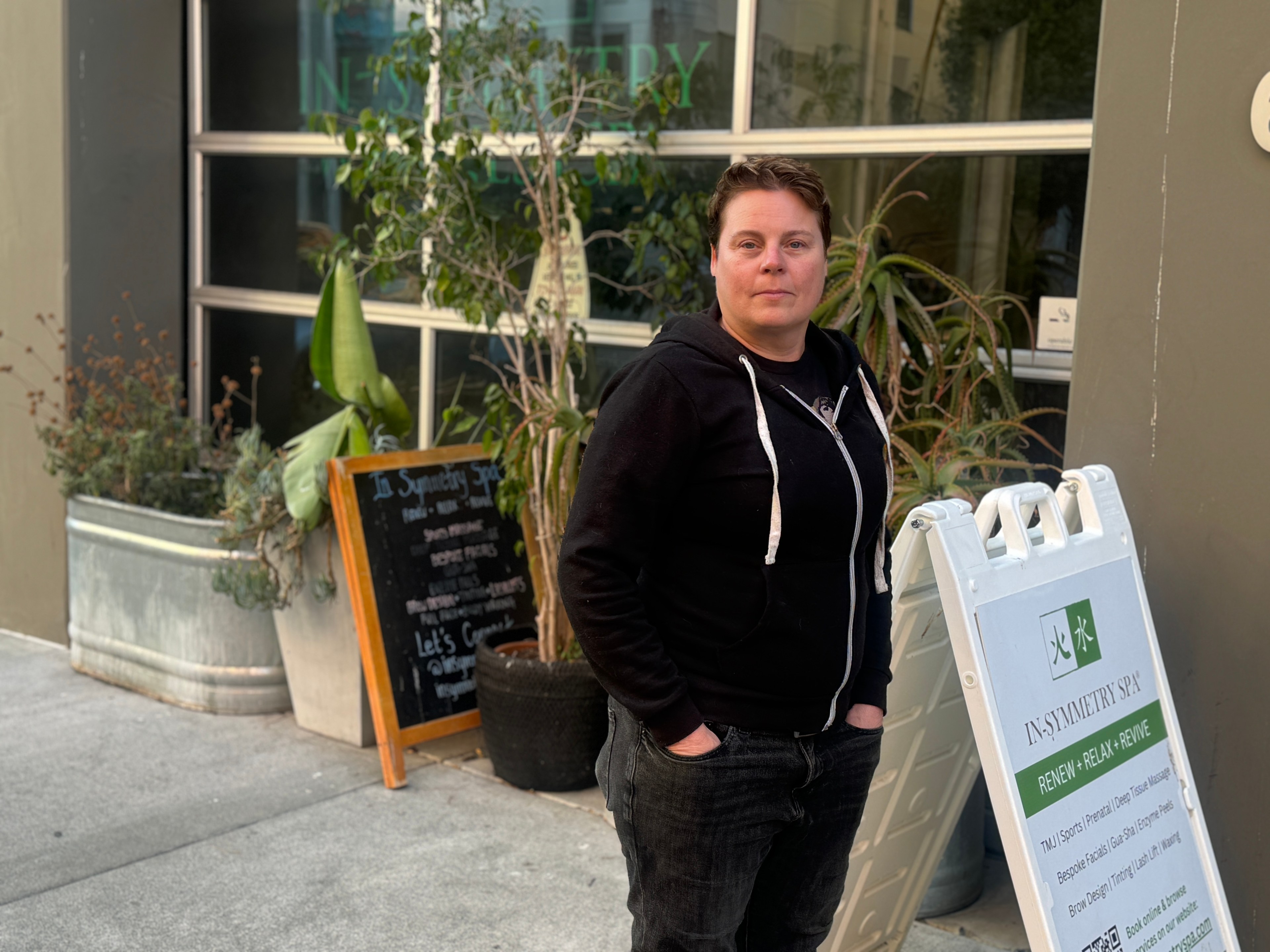 A person in a black hoodie stands outside a building with a sign for &quot;In-Symmetry Spa.&quot; There are potted plants and a chalkboard menu nearby.