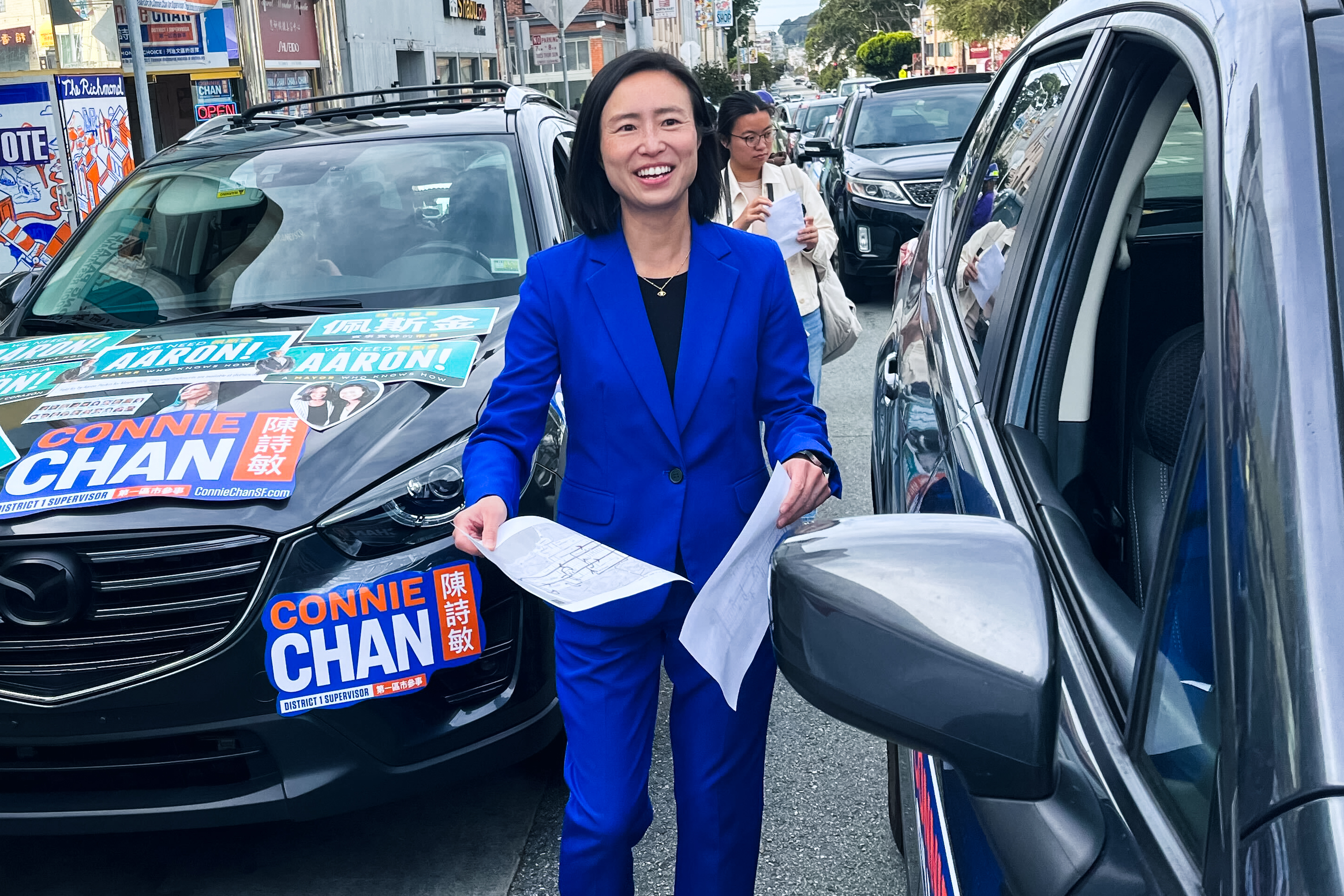 A person in a blue suit stands between two parked cars on a street. The car on the left is covered with campaign posters and signs. The person smiles while holding papers.