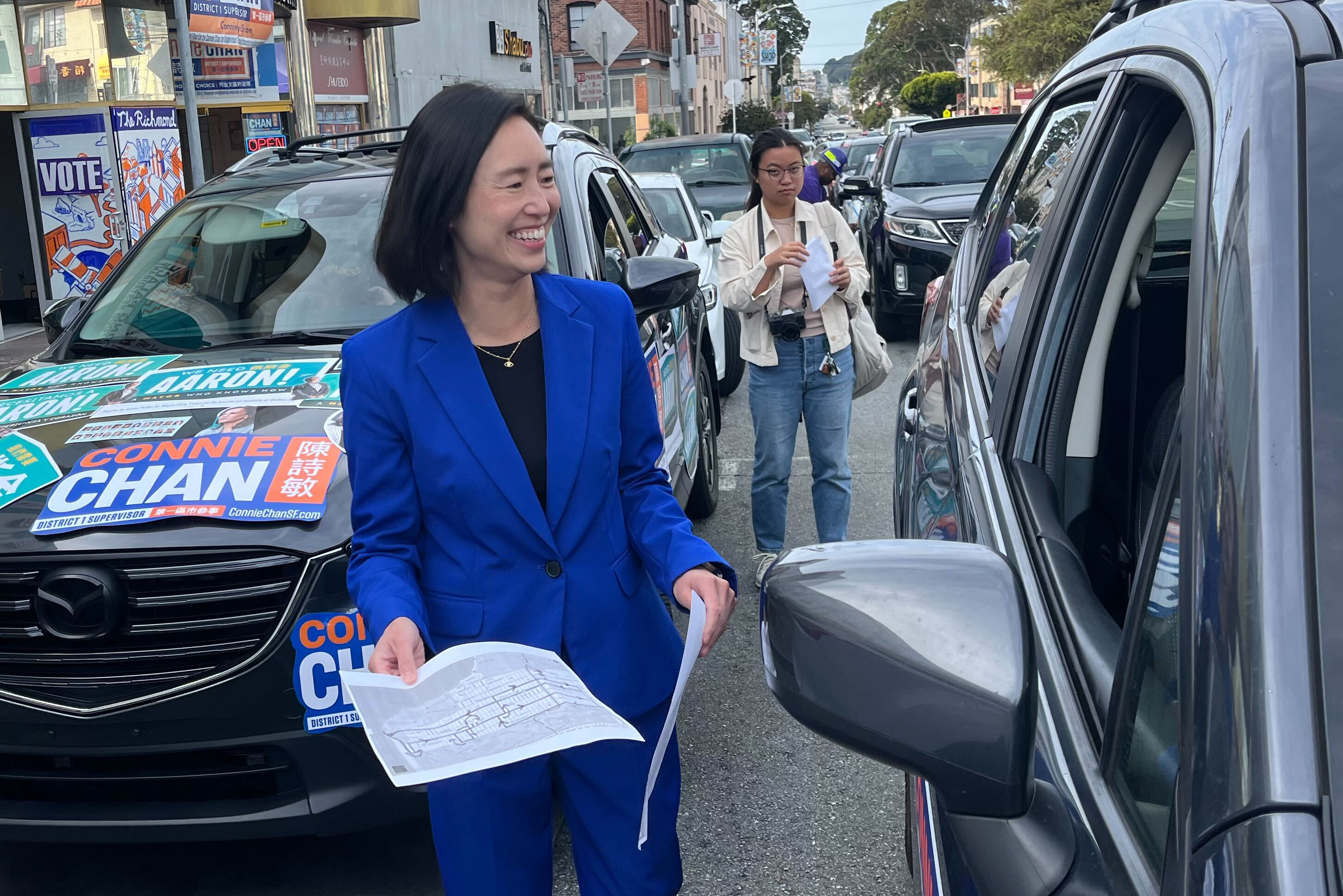 A woman in a blue suit smiles while holding papers by a car with campaign signs. Another person stands behind with a camera and paper, in an urban street setting.