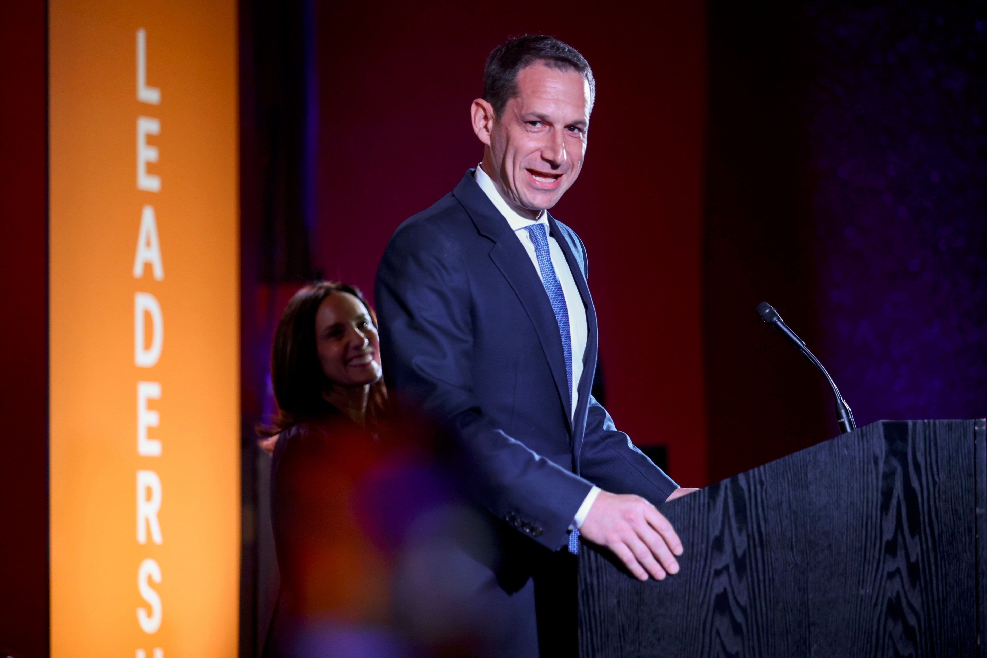 A man in a suit speaks at a podium with &quot;LEADERSHIP&quot; visible on a nearby orange background. A smiling woman stands behind him.