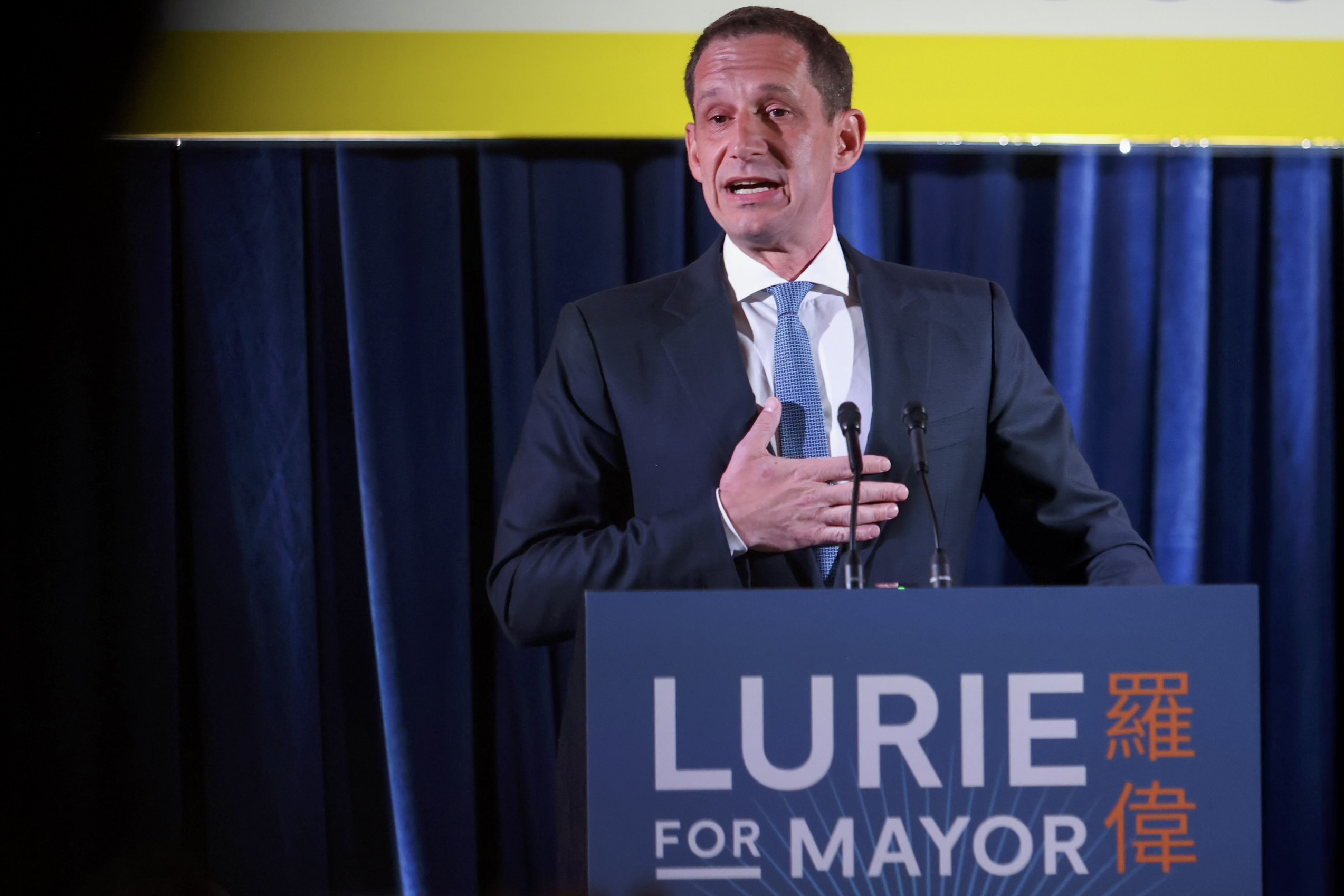 A man in a suit speaks at a podium with "Lurie for Mayor" written on it, standing in front of a blue curtain.