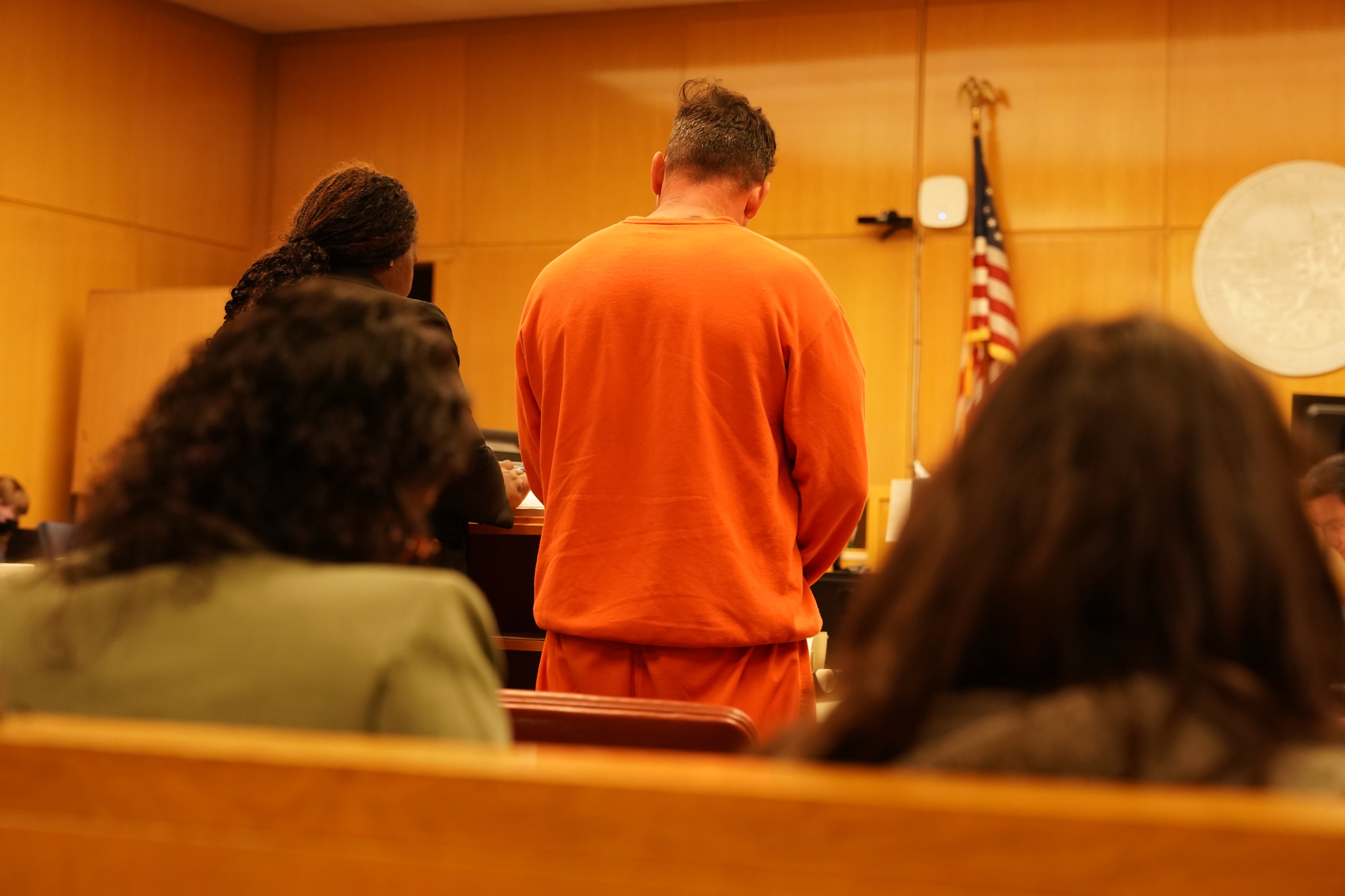 A person in an orange jumpsuit stands in a courtroom with their back to the camera. An American flag and the courtroom setting are visible.