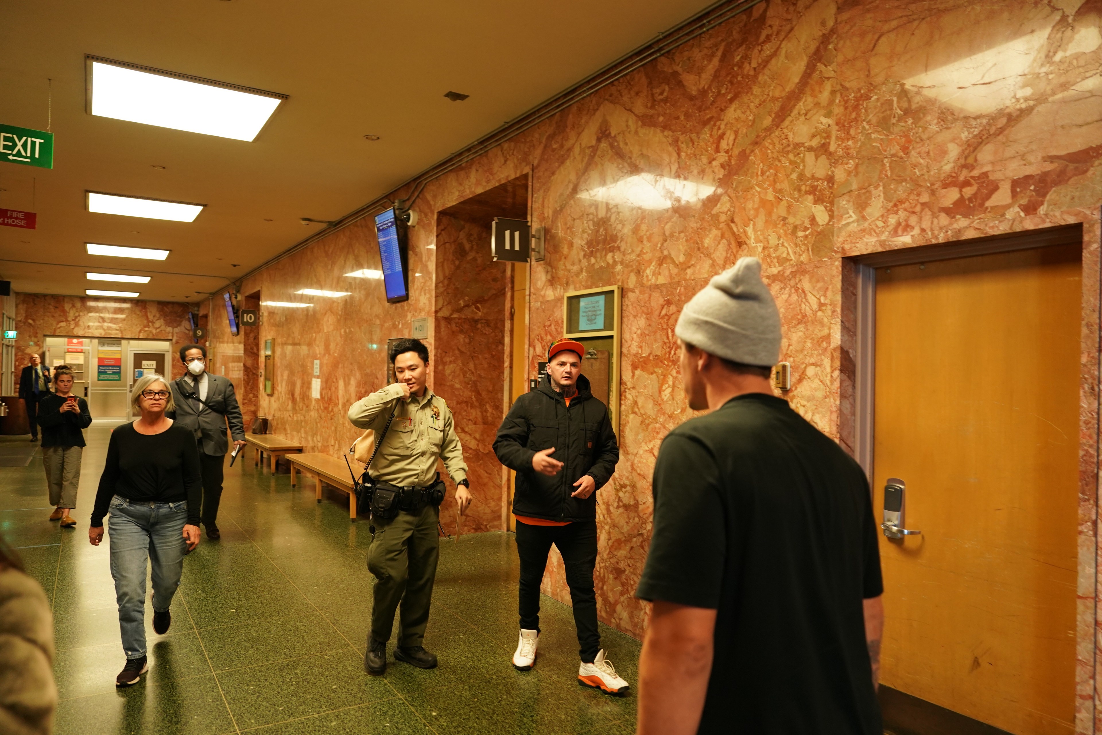 People walk through a hallway with marble walls and a green floor. One person is a uniformed officer, and others appear to be casually dressed. An exit sign is visible.