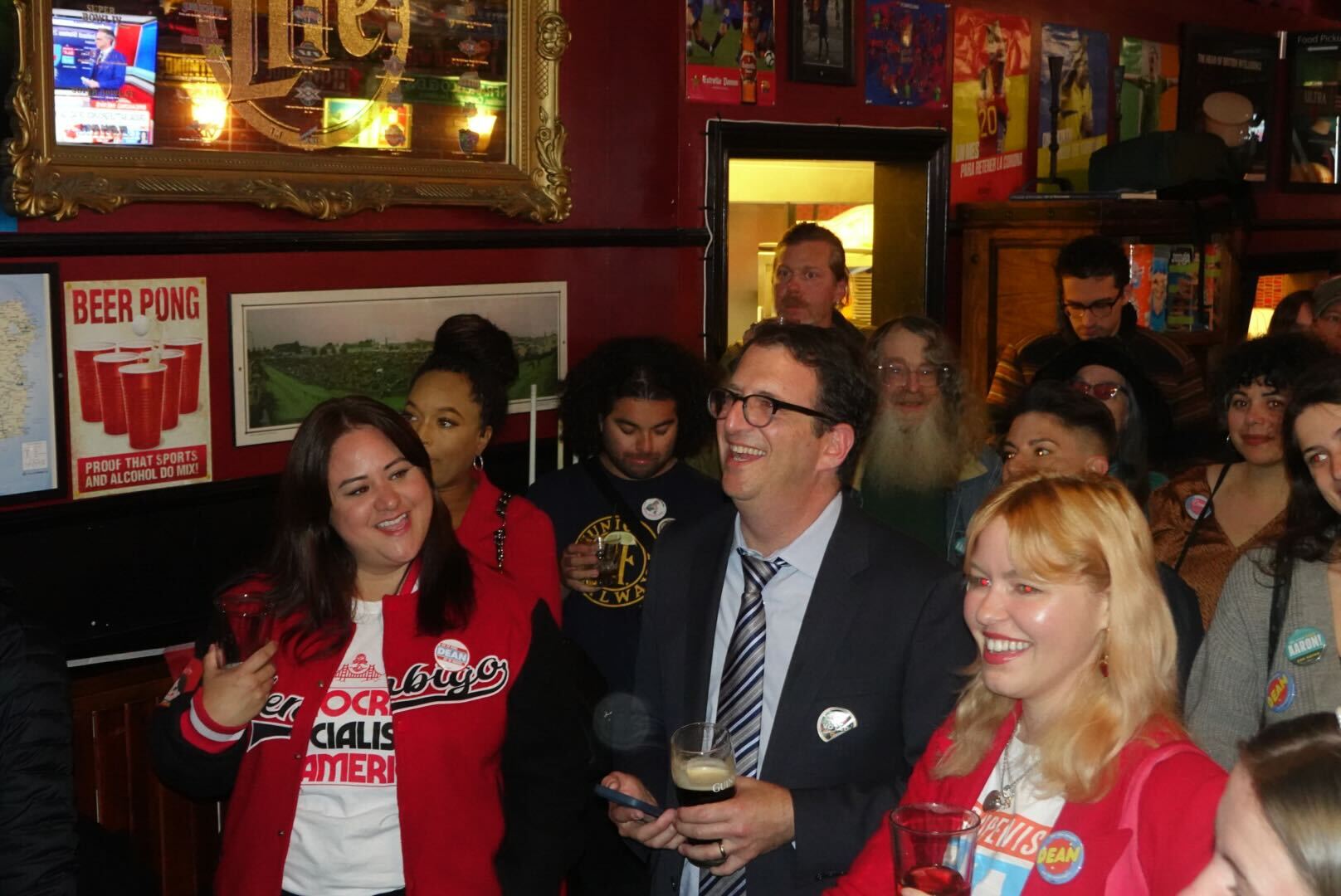 A group of people at a bar look happy and engaged, with several wearing campaign buttons. A man in a suit holds a drink and phone, enjoying the lively atmosphere.