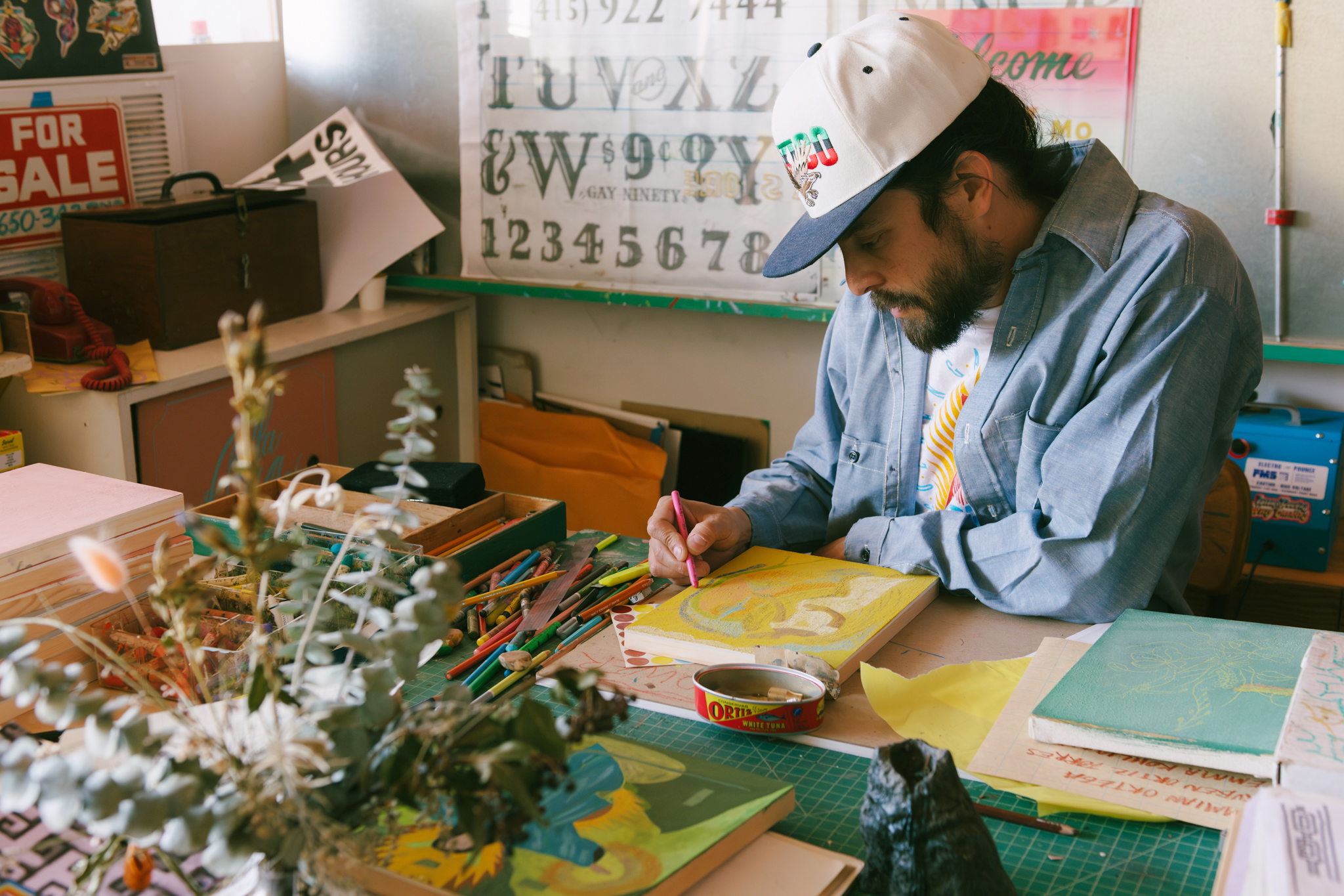 A man in a cap and denim shirt is drawing on a canvas at a cluttered table with pencils, papers, and plants. The workspace is filled with art supplies and signs.