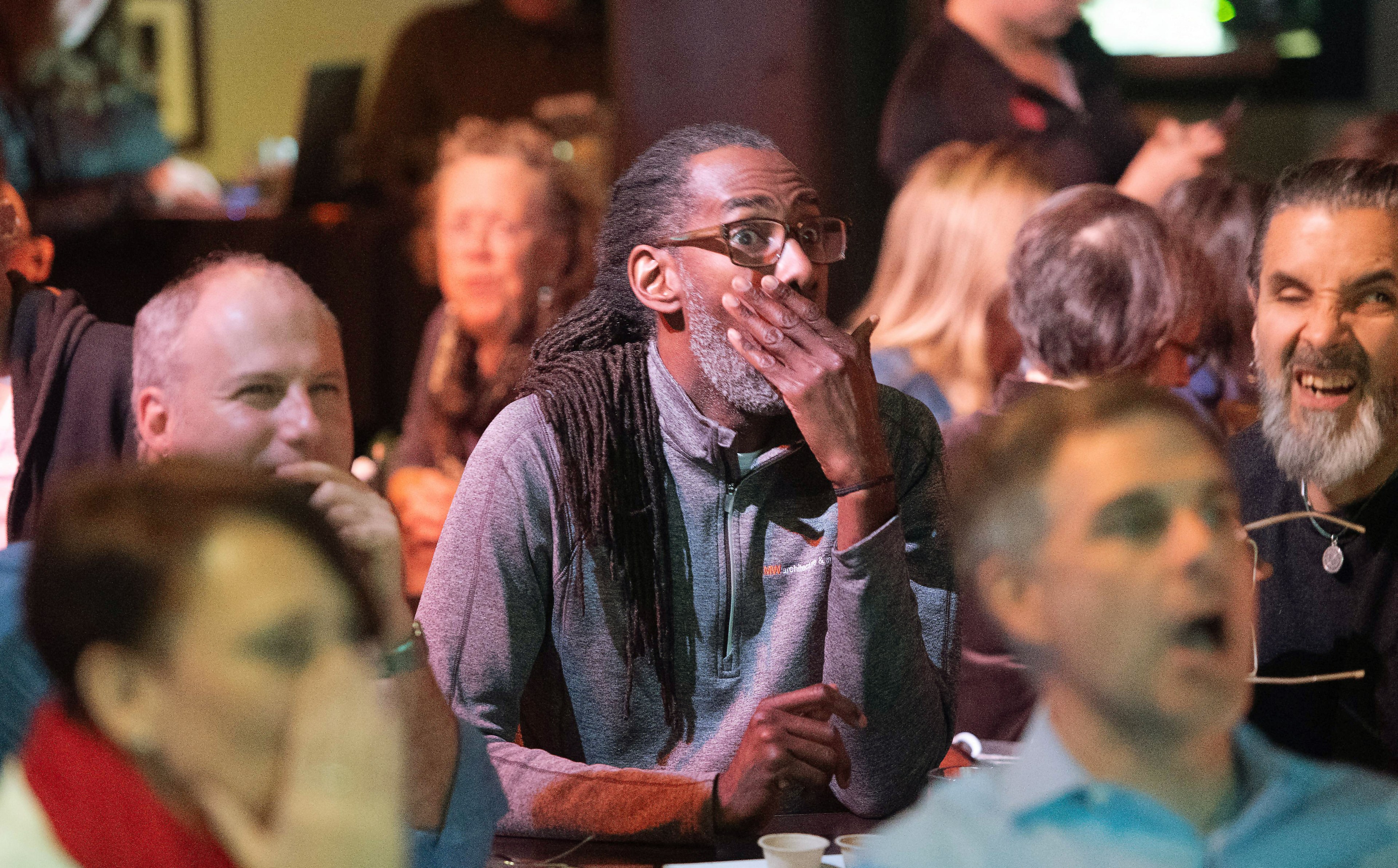 A group of people are indoors, appearing surprised or excited, with one person in the center holding their hand to their mouth as others around them react energetically.