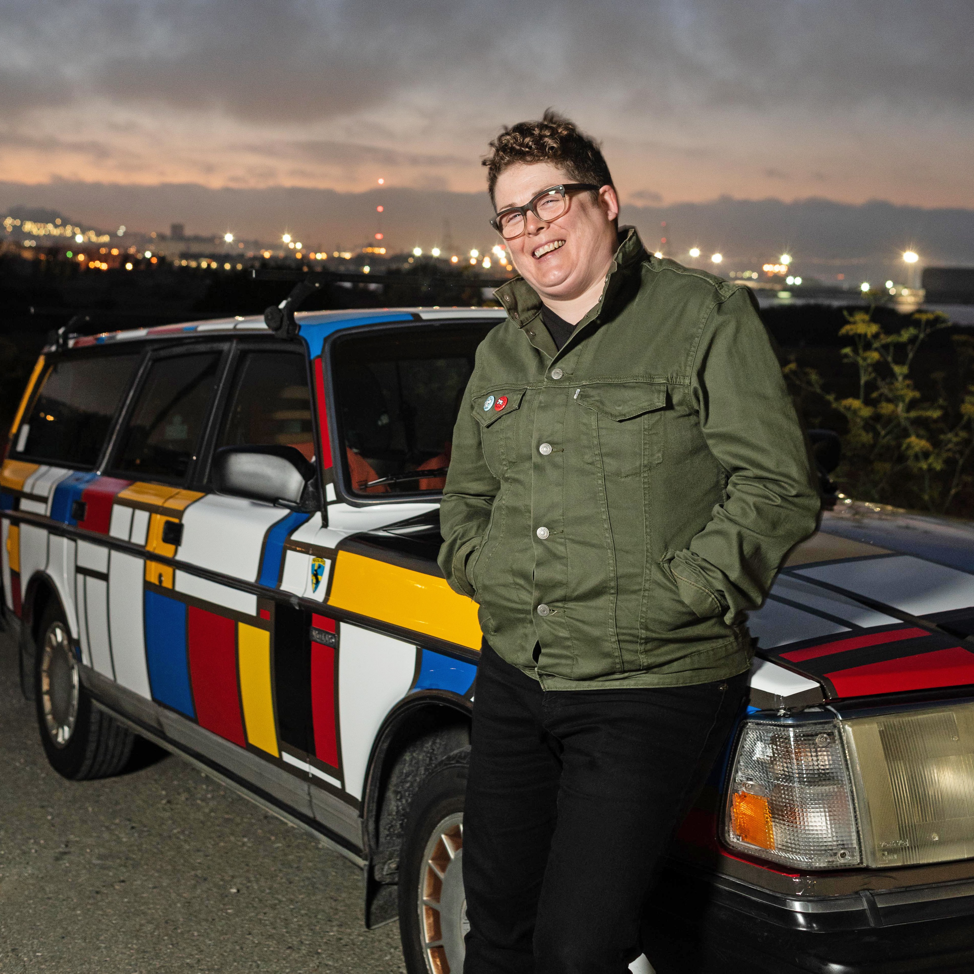 A person wearing glasses and a green jacket leans against a vibrantly patterned car with a geometric design of red, blue, yellow, and black lines at dusk.