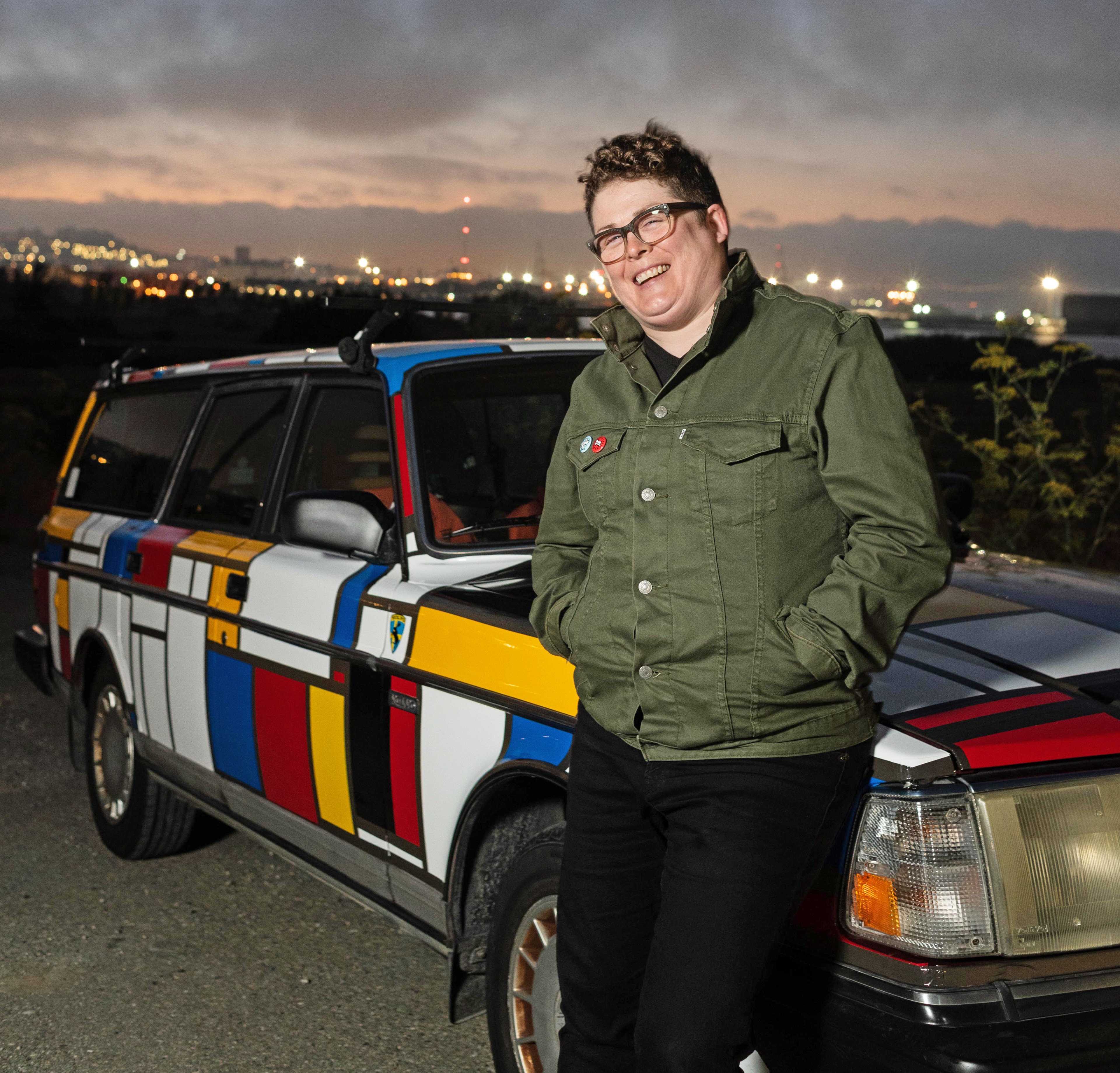 A person wearing glasses and a green jacket leans against a vibrantly patterned car with a geometric design of red, blue, yellow, and black lines at dusk.