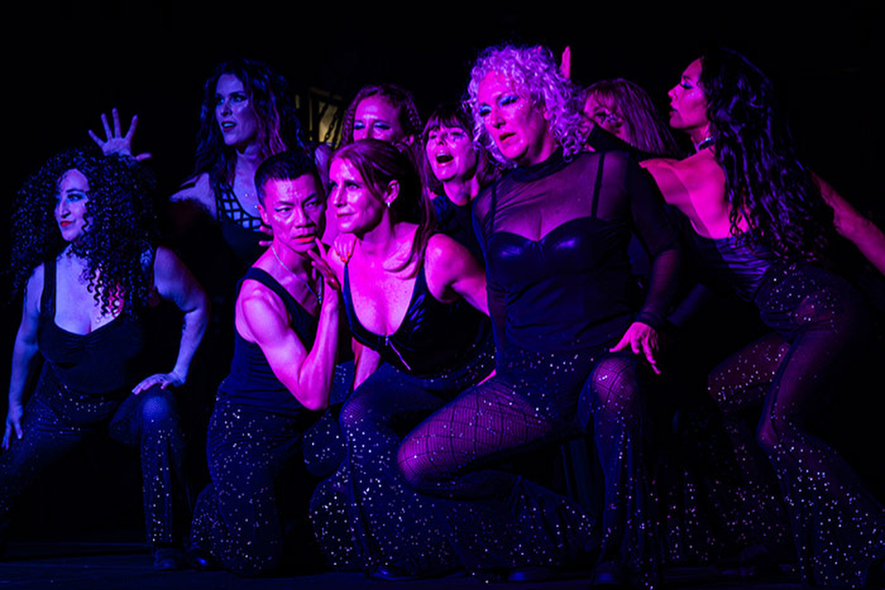 A dynamic group performance with nine people in dark, glittery costumes under purple lighting, striking expressive poses and conveying intense energy.